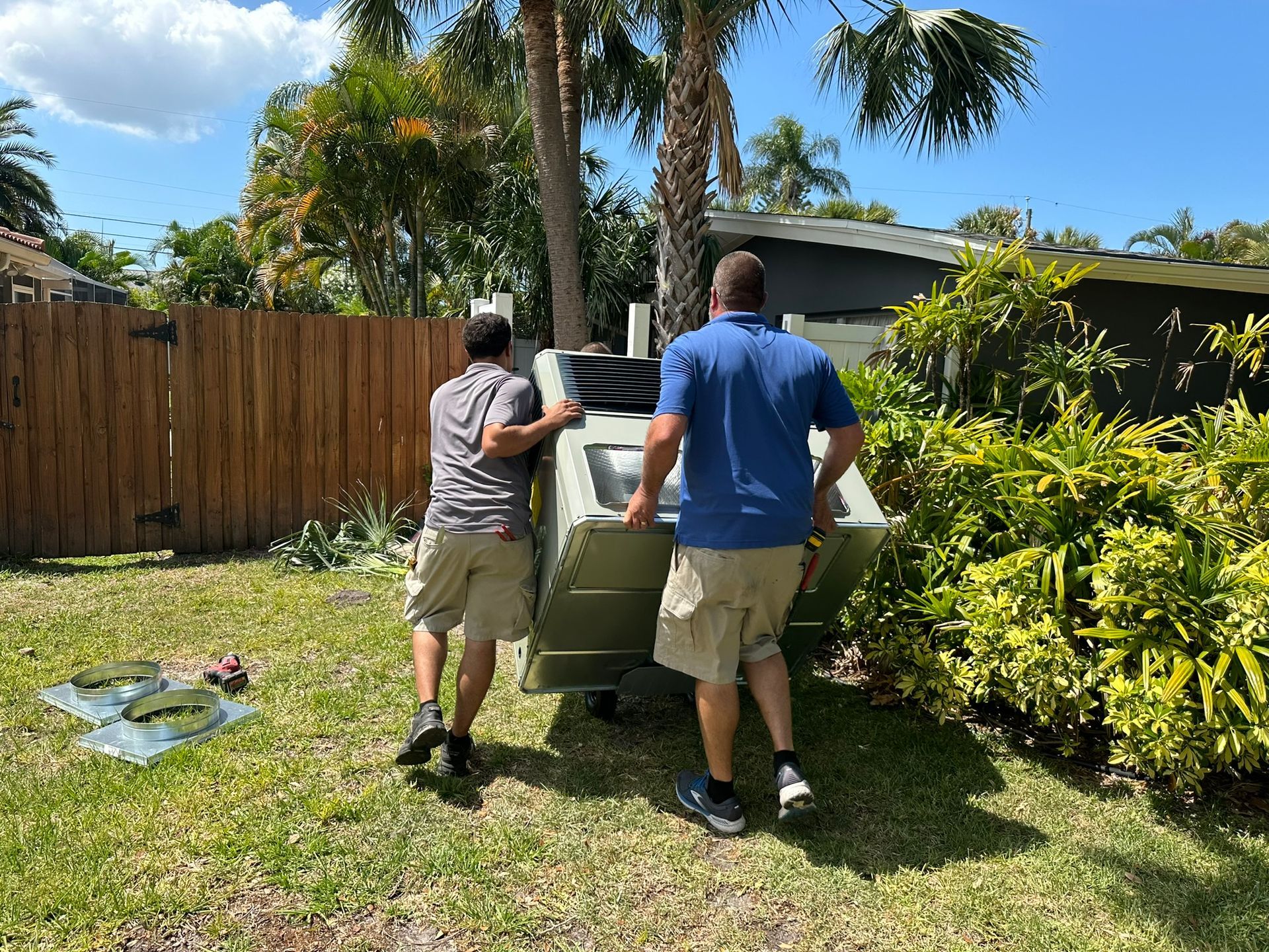 Two people carrying a large appliance across a grassy yard with palm trees and a wooden fence.