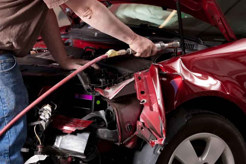Mechanic repairs a damaged red car with tools in a shop.