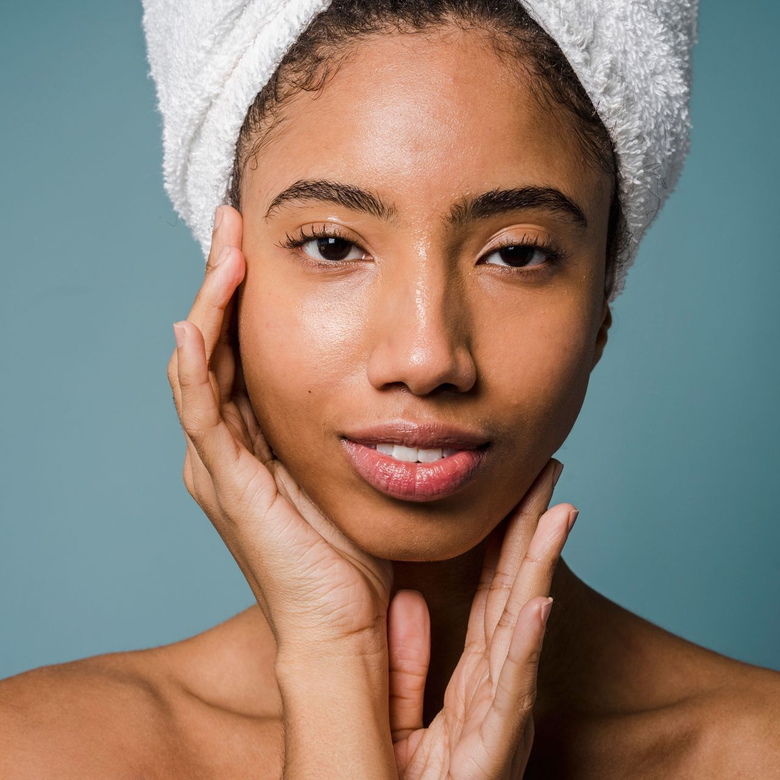 Woman with towel wrapped around hair, gently touching her face against a blue background