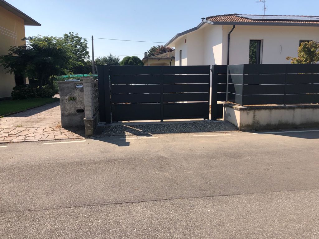 Un cancello a stecche orizzontali grigio scuro si apre su un vialetto asfaltato. Sono visibili la strada e una casa.