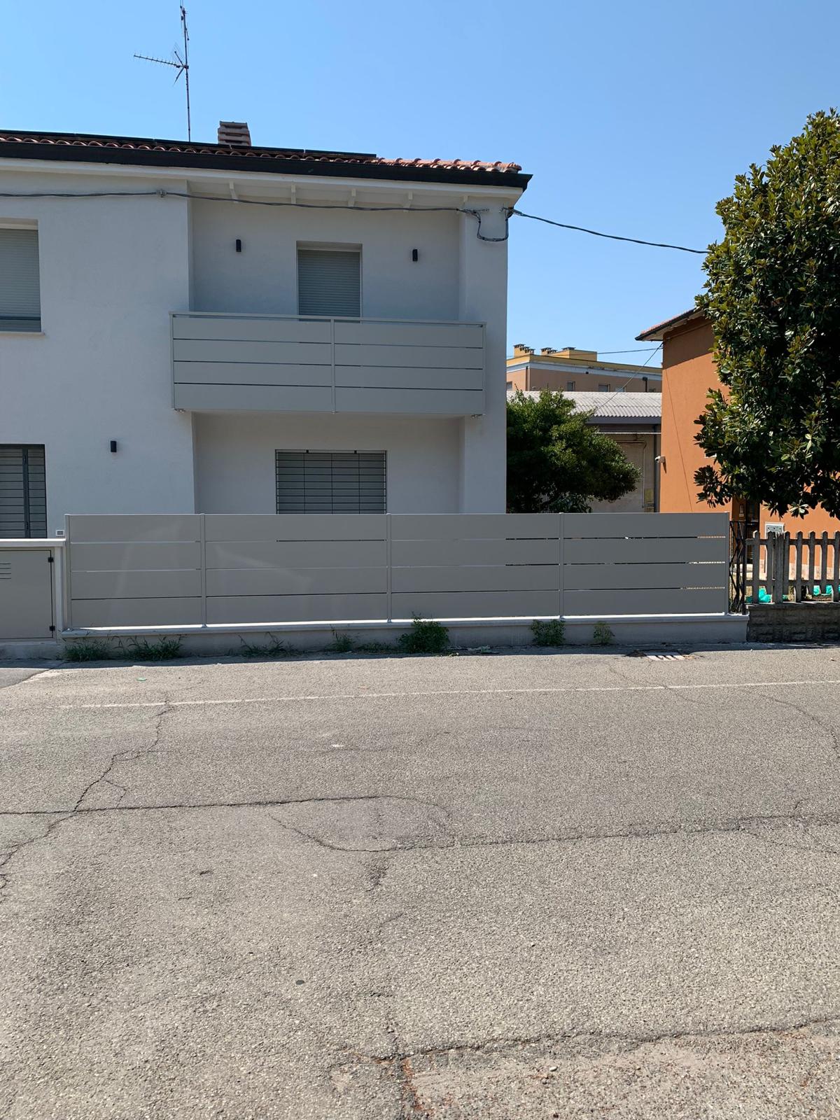 Edificio bianco a due piani con recinzione e balcone grigi. Un albero sulla destra e il cielo è azzurro.