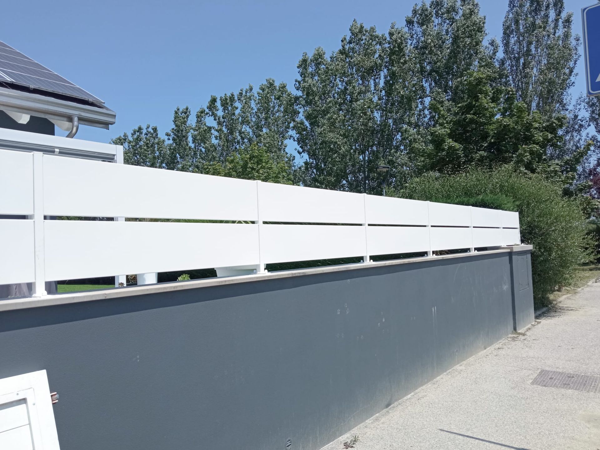 Recinzione bianca con stecche orizzontali in cima a un muro grigio, accanto a un marciapiede e ad alcuni cespugli verdi.