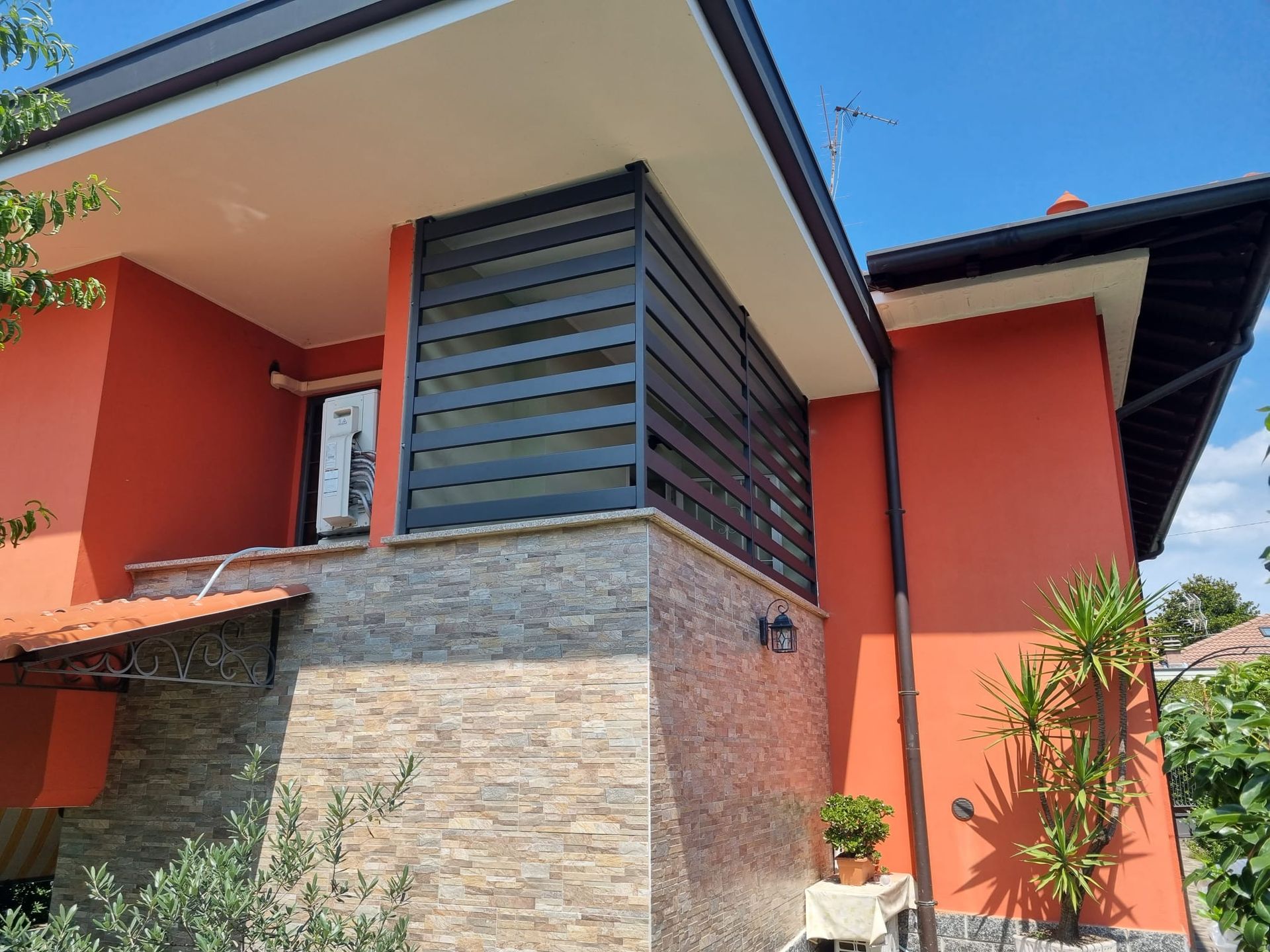 Casa arancione con facciata in pietra, balcone con listelli scuri, soffitto bianco e cielo azzurro.