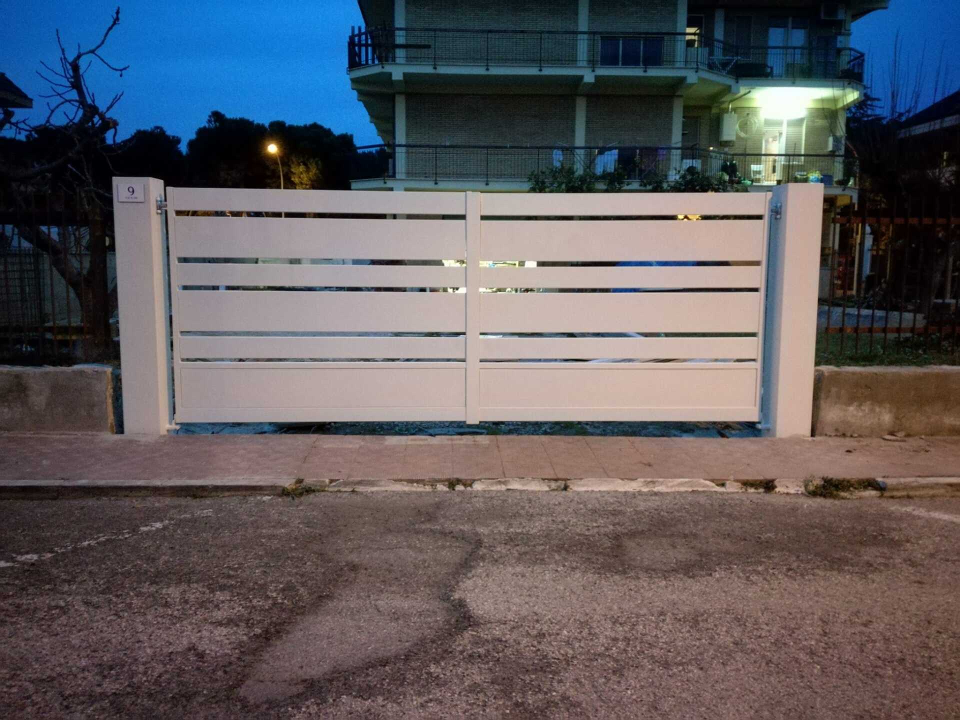 Cancello bianco con listelli orizzontali tra due pali di cemento, di fronte a un edificio, su una strada.