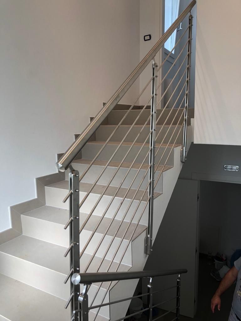 Scala in acciaio inossidabile con ringhiera in cavo, che sale contro una parete grigio chiaro.