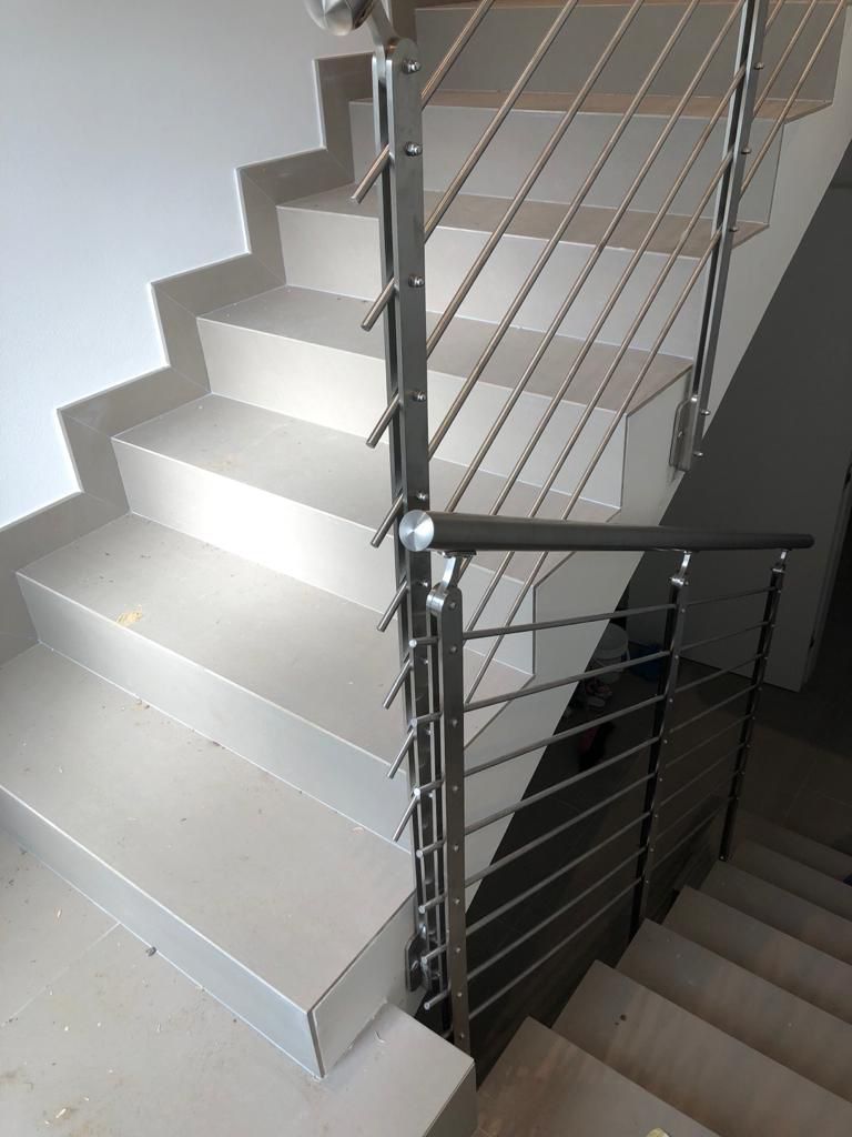 Scala con ringhiera in acciaio inox e corrimano scuro. Gradini grigi. Pareti bianche.