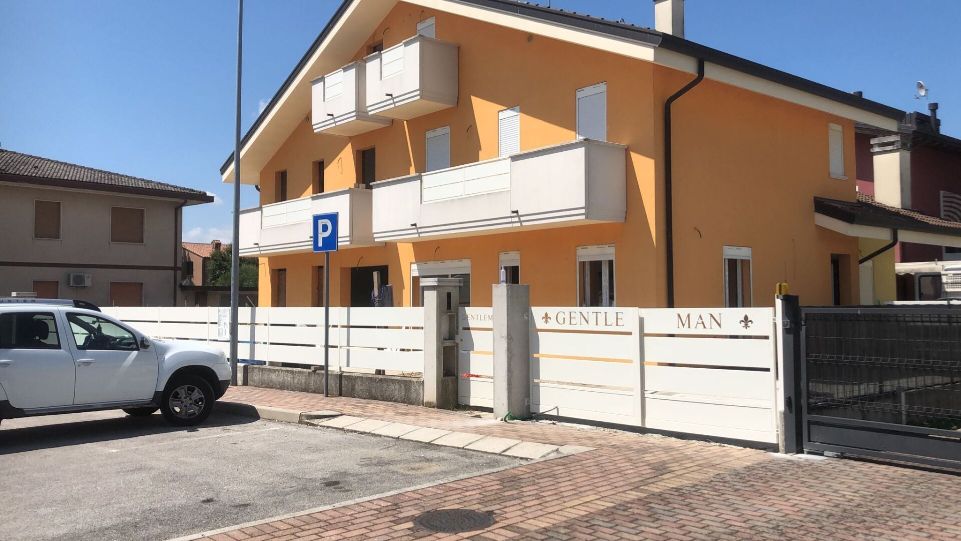 Casa gialla con recinzione bianca, balconi e cartello di parcheggio. SUV bianco parcheggiato di fronte.