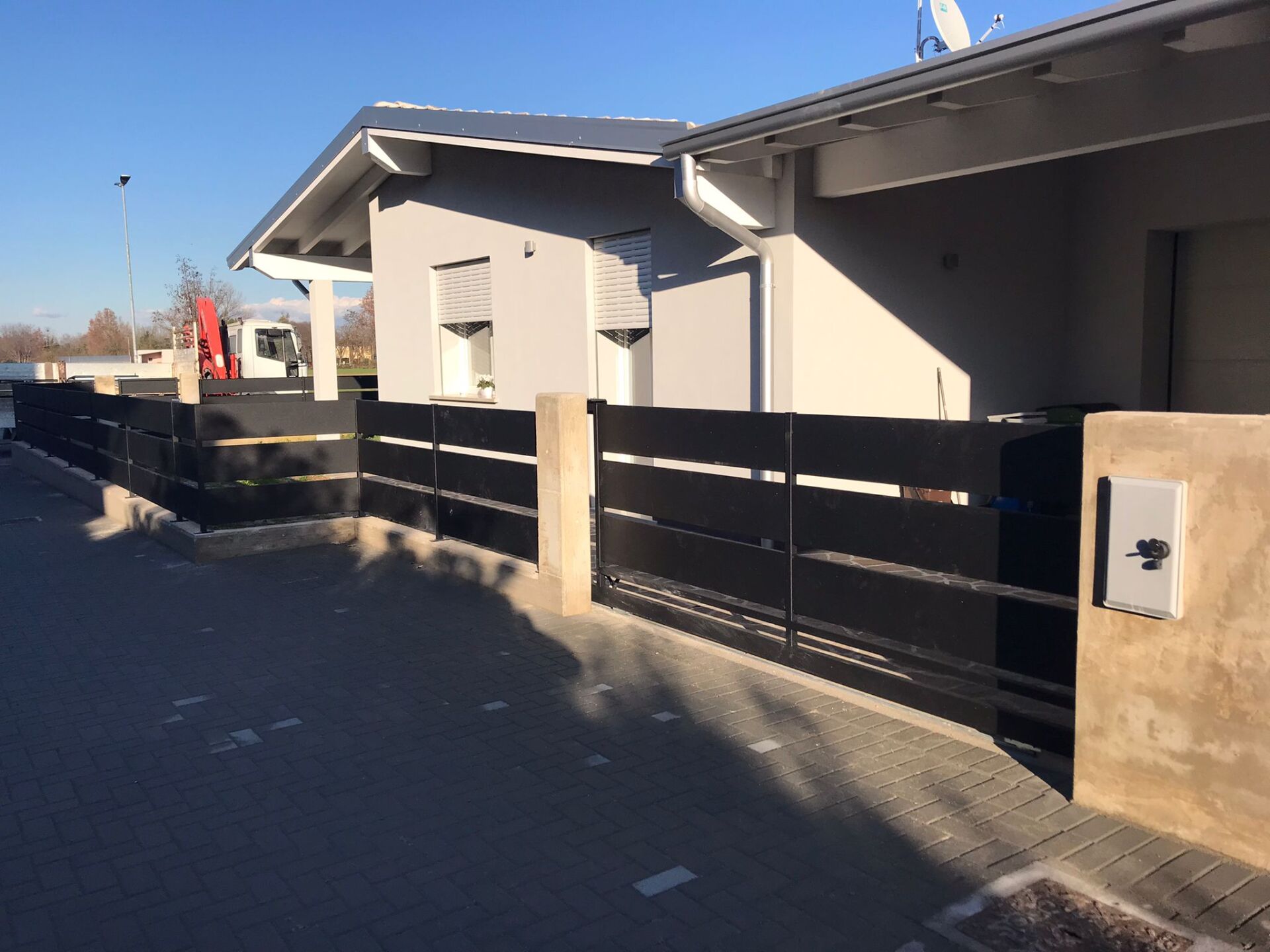 Recinzione moderna nera davanti a una casa chiara con vialetto pavimentato in una giornata di sole.