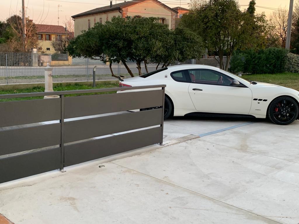 Una Maserati bianca parcheggiata dietro una recinzione grigia orizzontale su un vialetto di cemento.