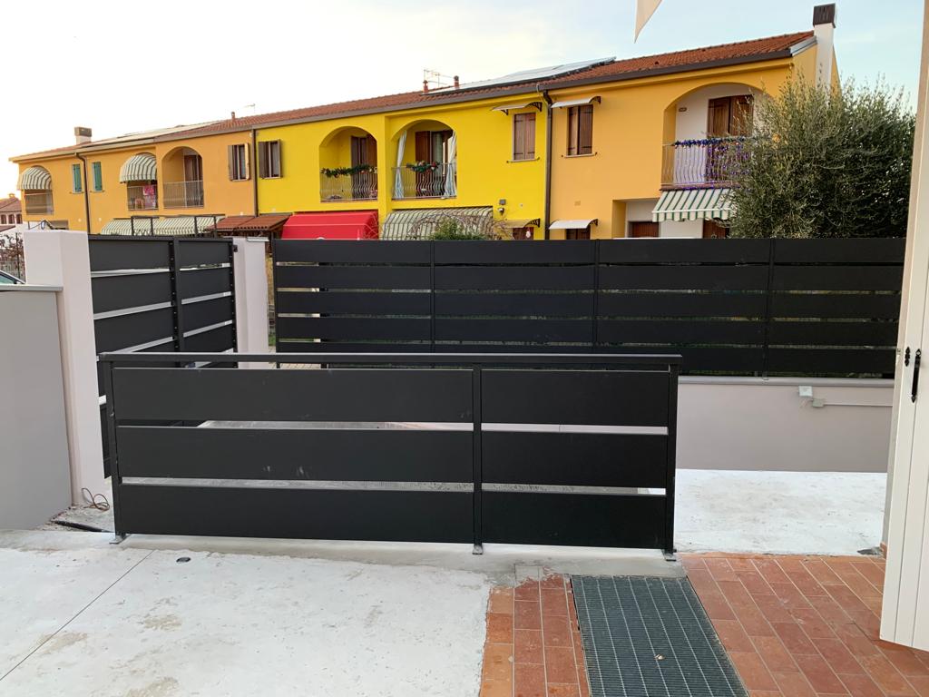 Recinzione e cancello con listelli orizzontali neri di fronte a edifici gialli e beige.