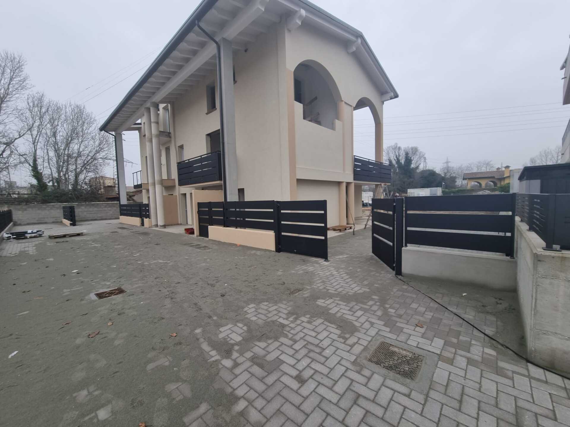 Edificio beige a due piani con recinzione orizzontale nera e cancello, giornata nuvolosa.