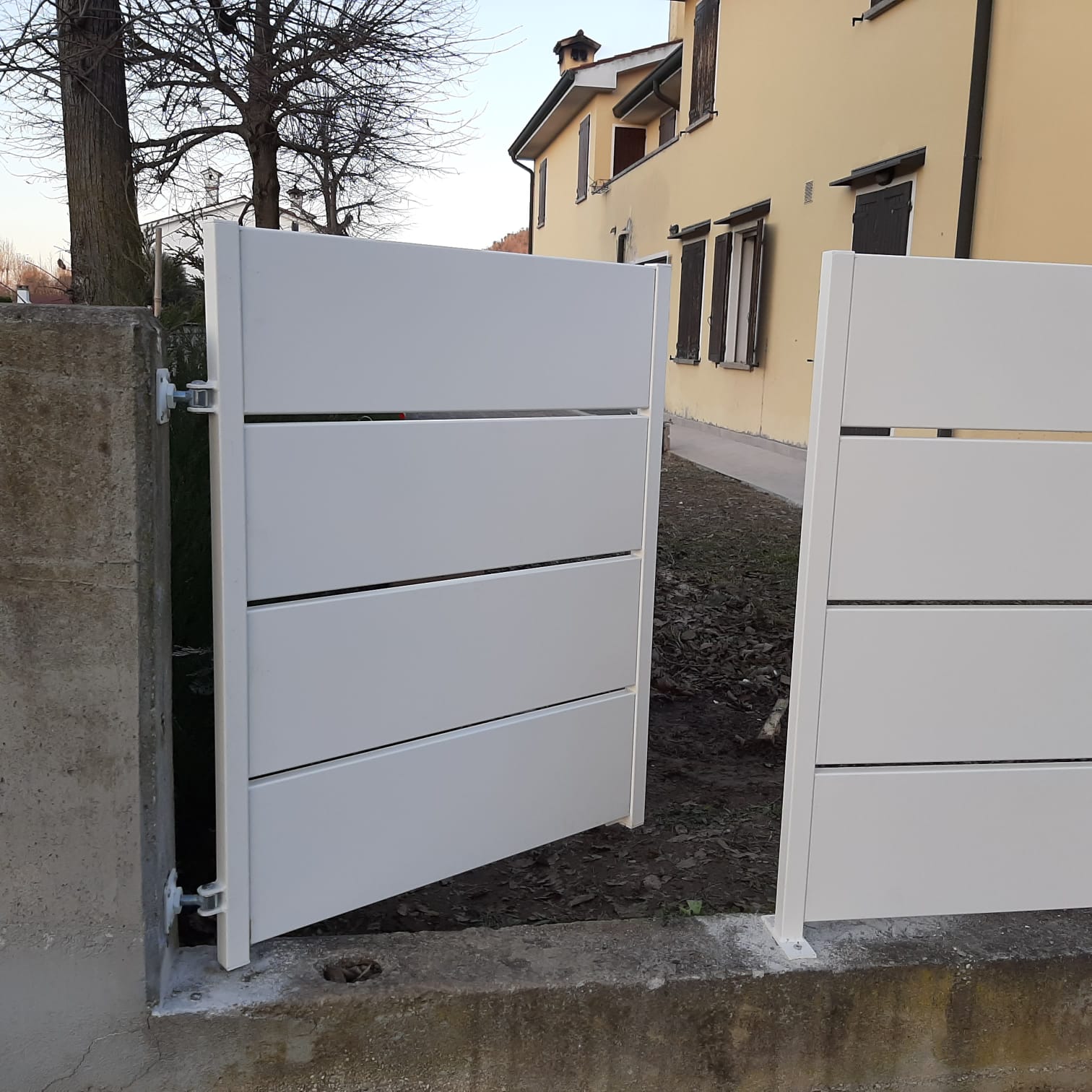 Cancello bianco, parzialmente aperto, attaccato a un muro di cemento. Area recintata vicino a una casa gialla.