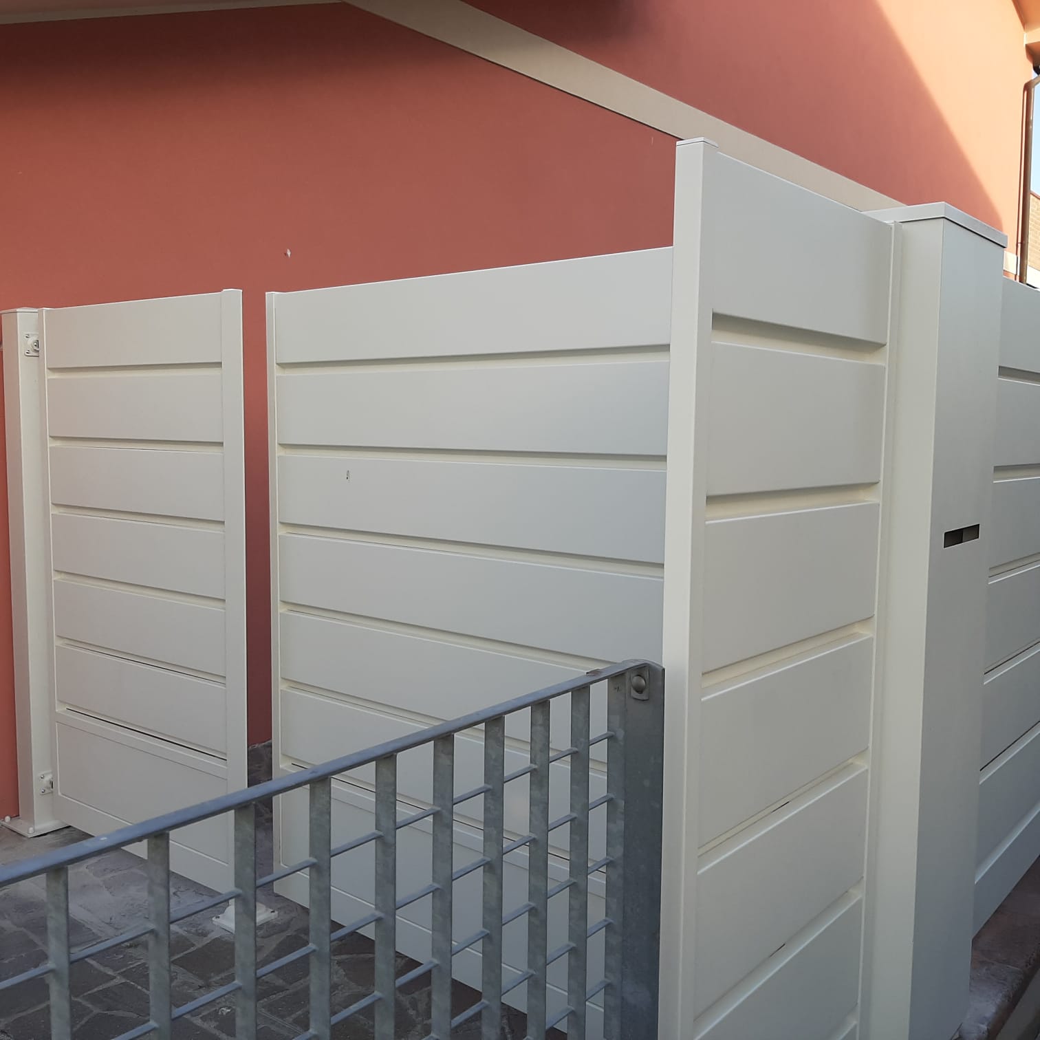 Cancelli e recinzione con stecche bianche contro un muro rosso, accanto a una ringhiera metallica.