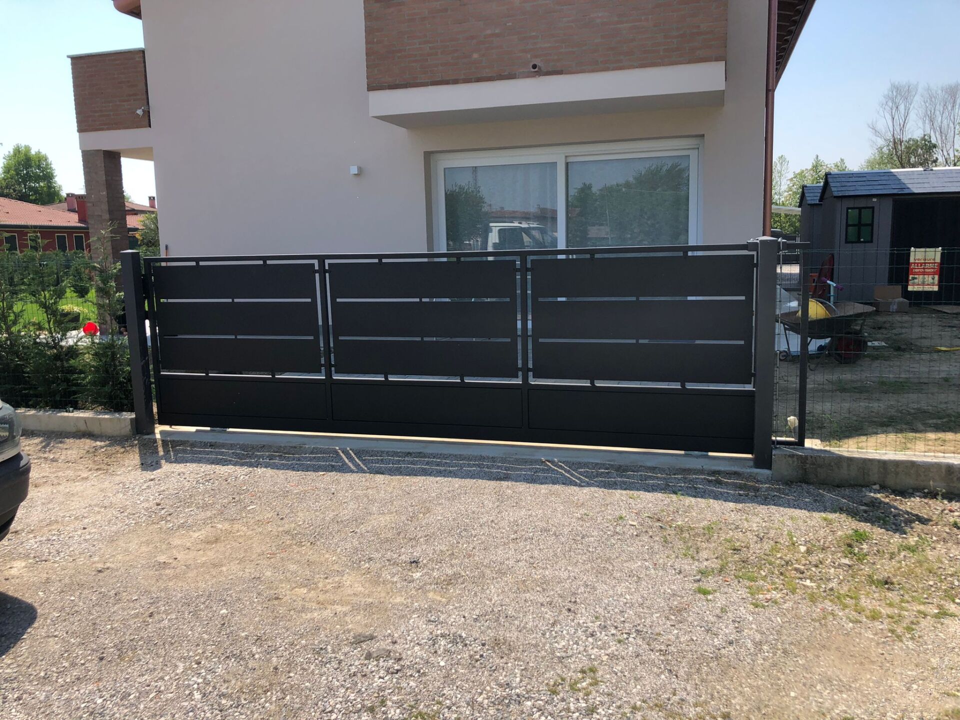 Cancello di metallo nero davanti a una casa chiara. Vialetto di ghiaia in primo piano.