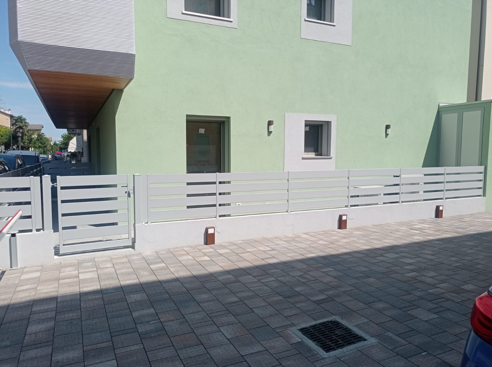 Edificio verde chiaro con recinzione grigia e cortile pavimentato.