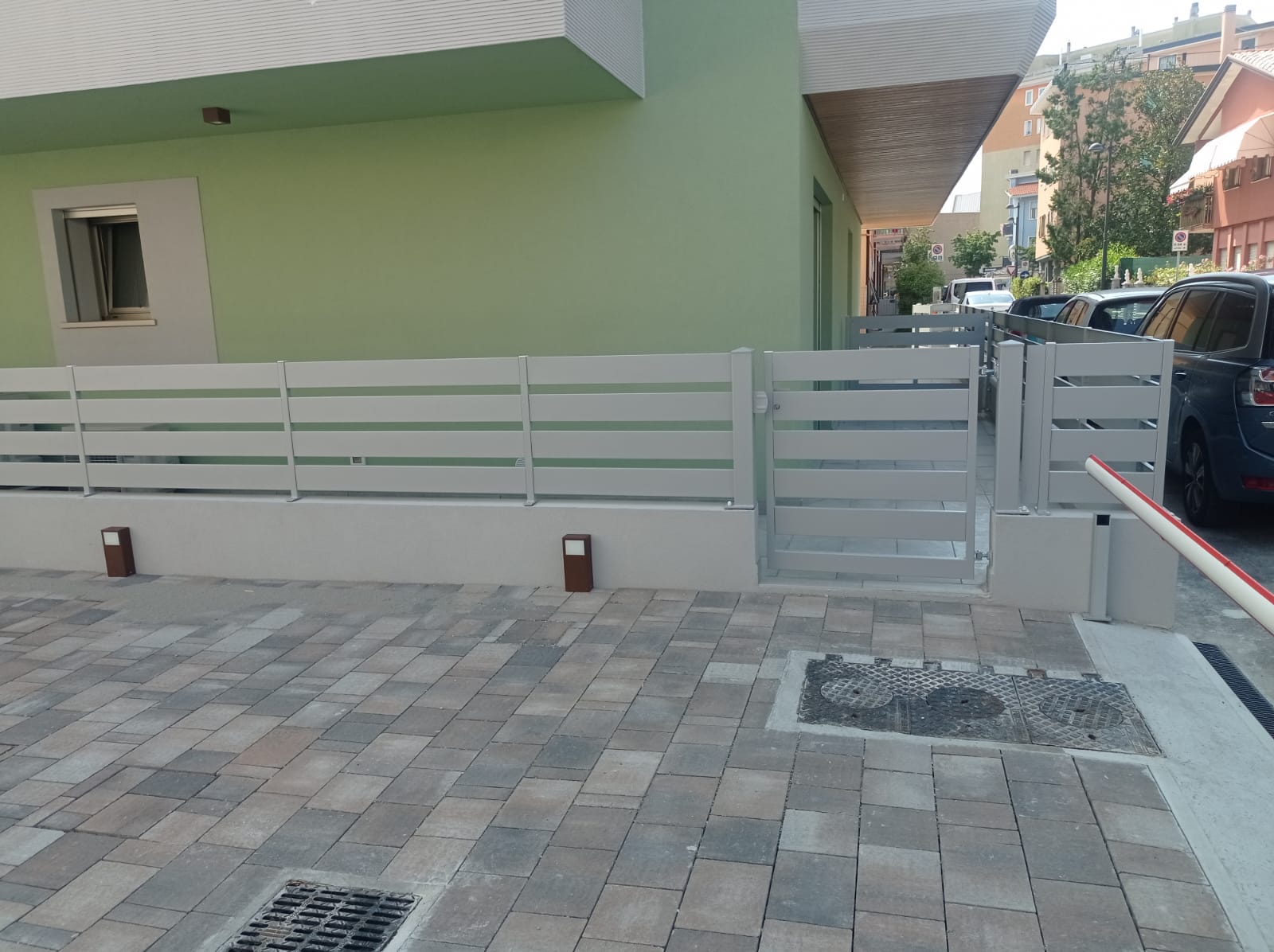 Una recinzione e un cancello grigio chiaro delimitano un'area pavimentata di fronte a un edificio verde chiaro.