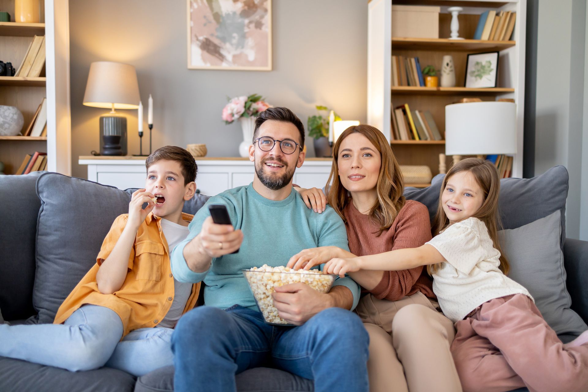 A family is sitting on a couch watching tv and eating popcorn.