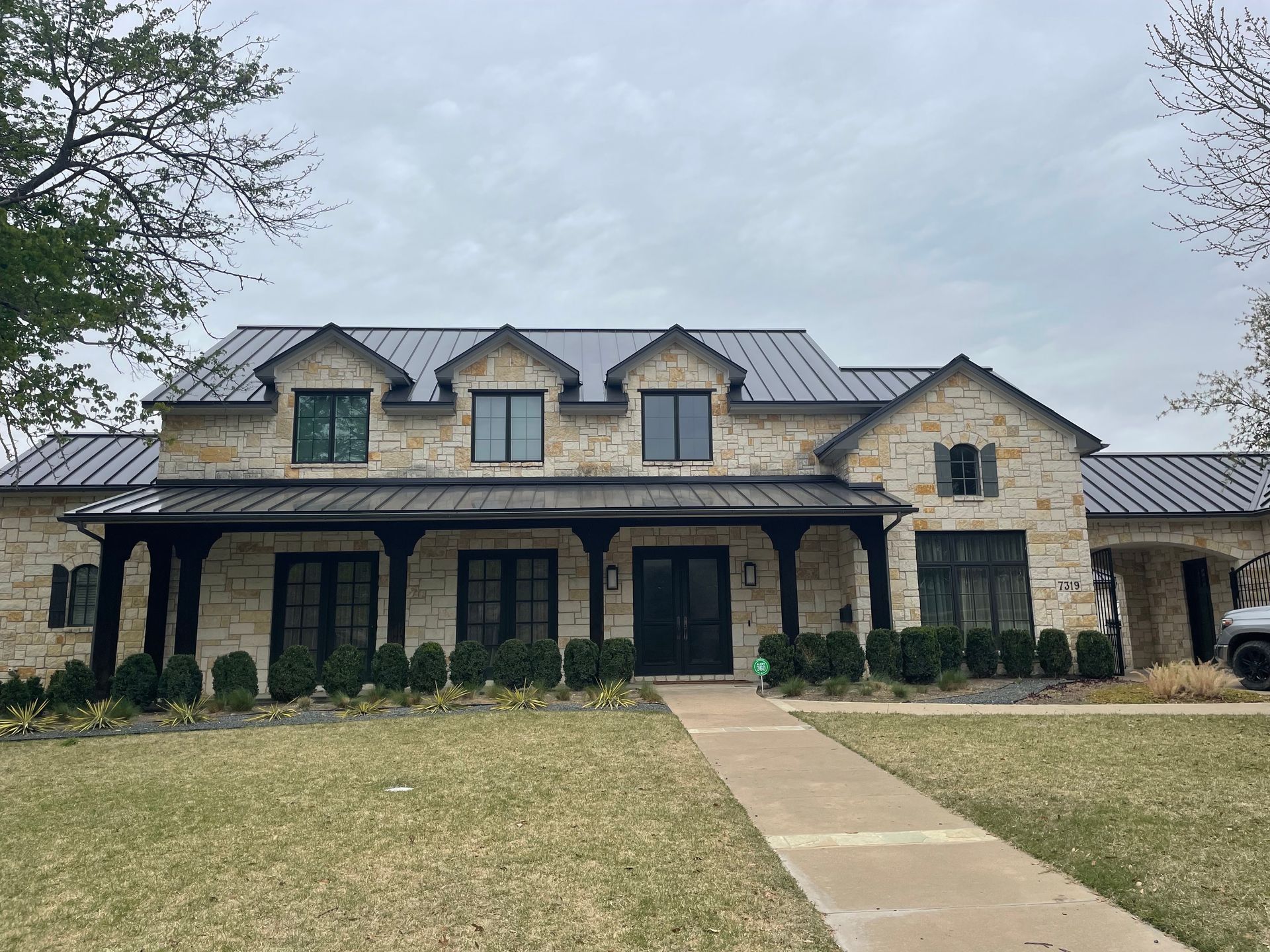 Stone two-story house with dark roof, arched porch, and front walkway under cloudy sky