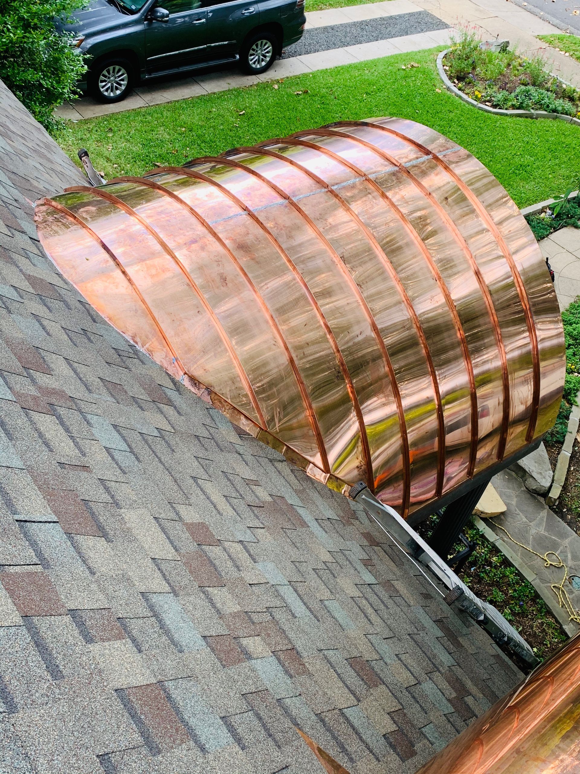 Copper-colored arched roof extension on a house beside a shingled roof and driveway.