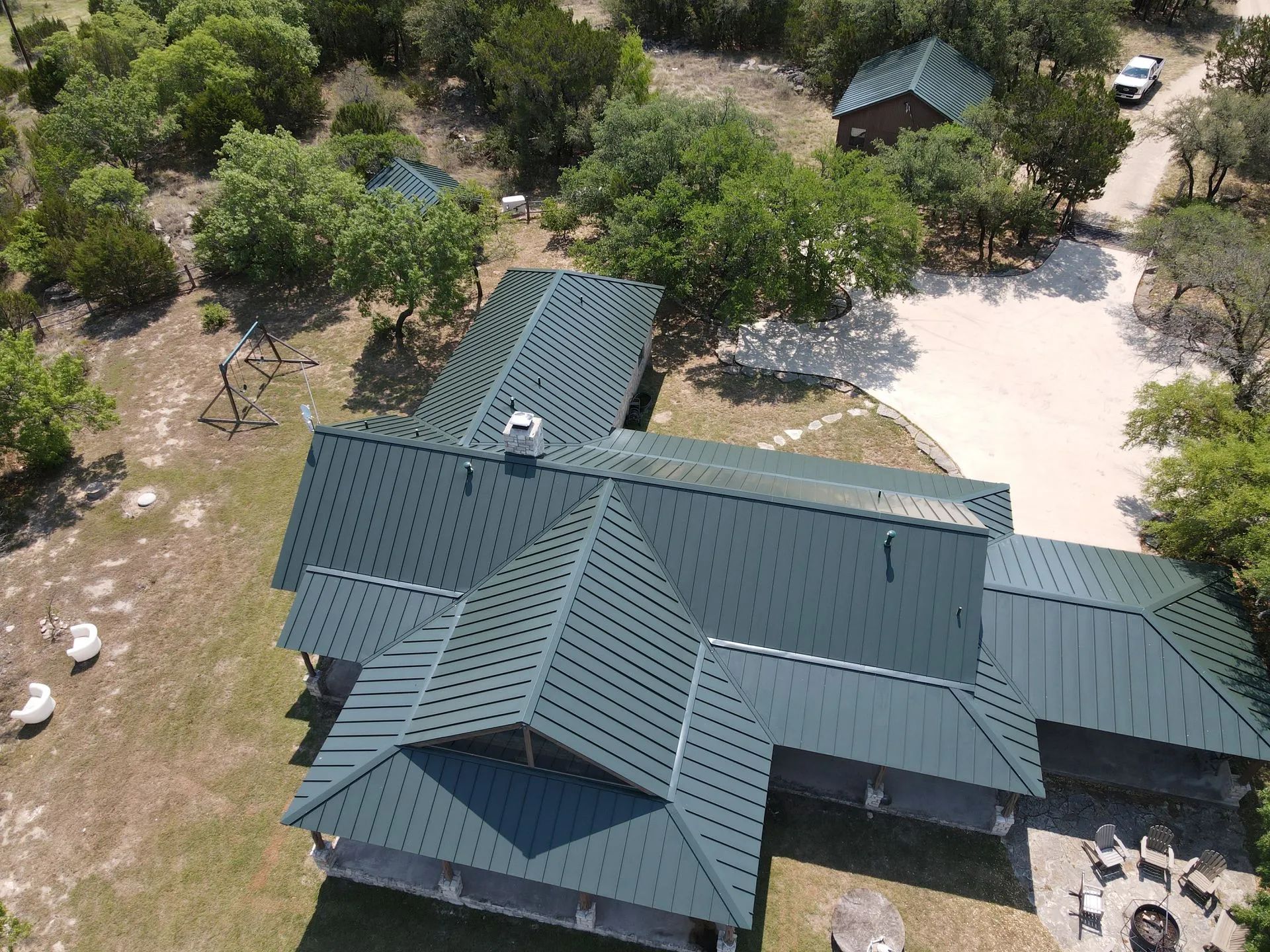 Aerial view of a large house with a dark green roof surrounded by trees and a dirt driveway.