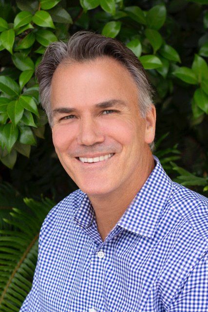 Smiling man in a blue checkered shirt standing in front of green foliage