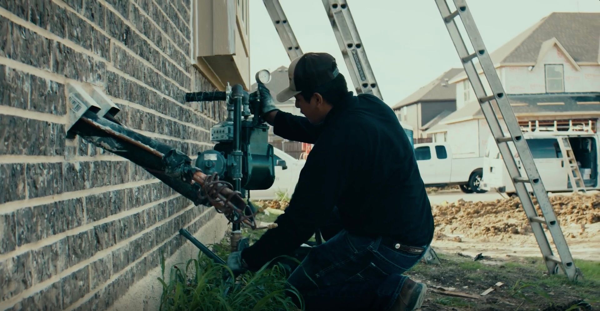 Worker using a nail gun beside a brick wall during house construction, with ladders in the background.