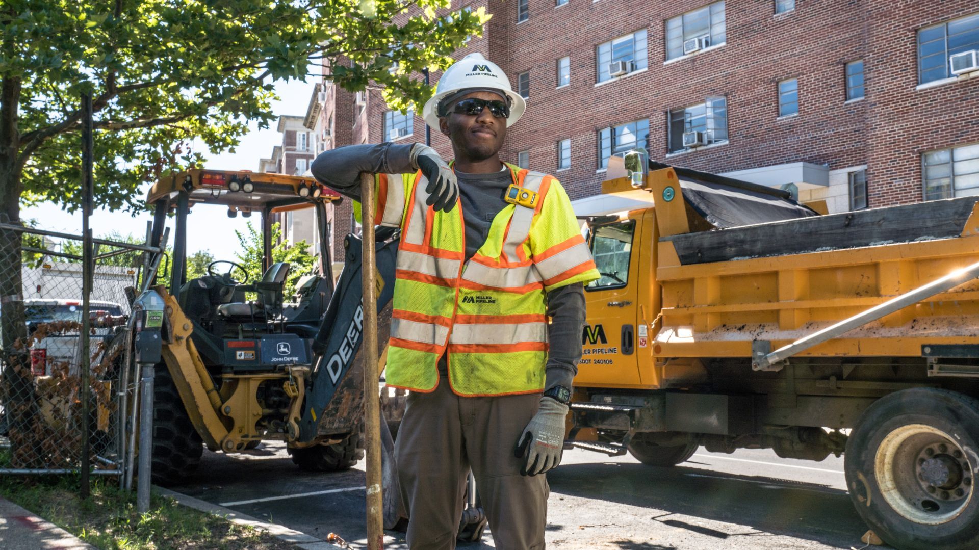 Construction worker in reflective vest and hard hat stands beside a backhoe and dump truck on a city street.