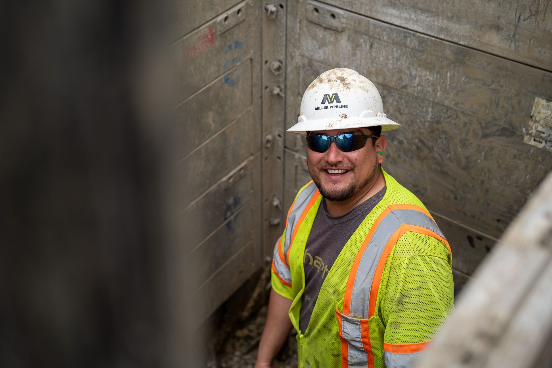 Construction worker in hard hat and safety vest standing in a trench, smiling at the camera.