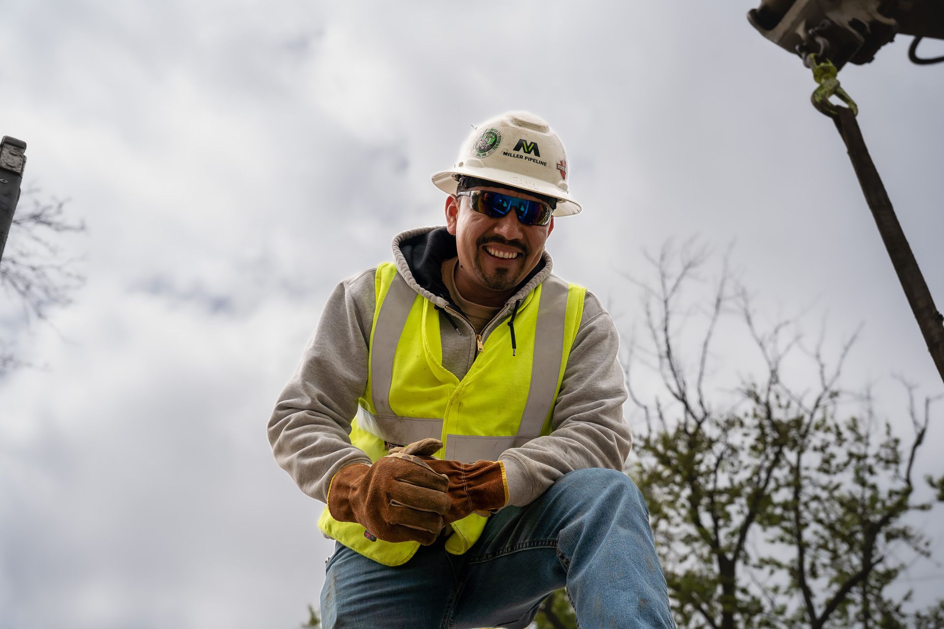 Worker in a hard hat and neon safety vest, crouching outdoors and smiling while holding a tool.