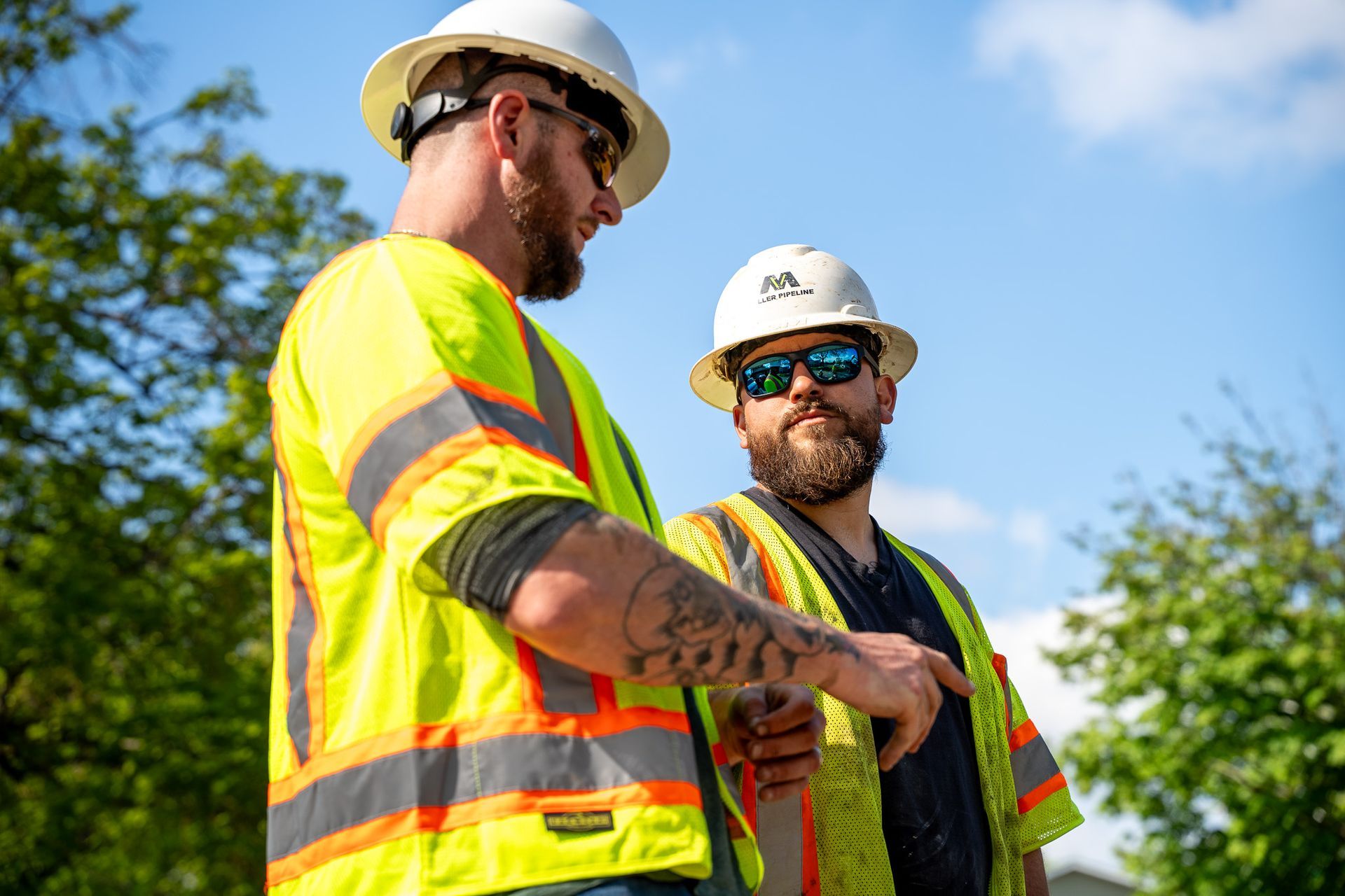 Two construction workers in safety vests and hard hats standing outdoors against a blue sky