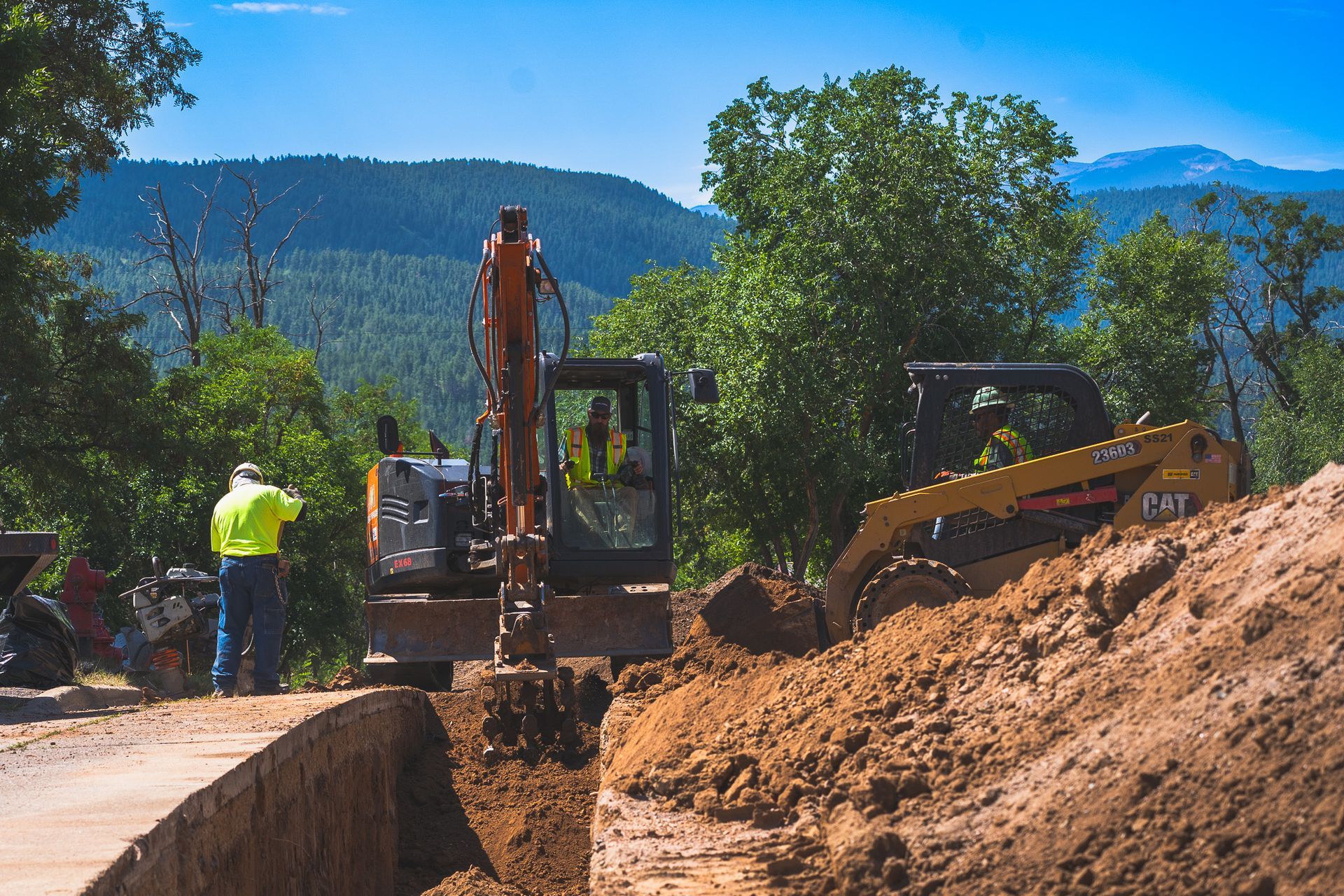 Construction crew digging trench with excavator on a dirt road, mountains and trees in the background