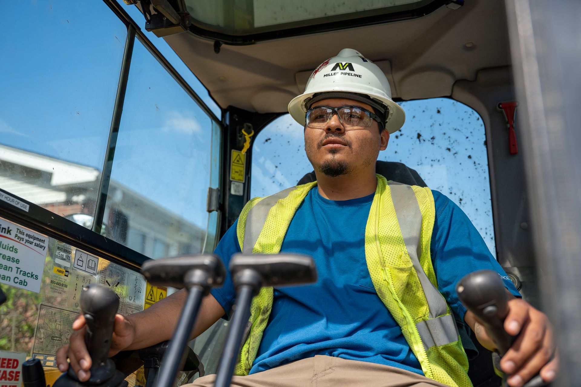 Construction equipment operator wearing a hard hat and safety vest, seated in a cab and gripping two joysticks.