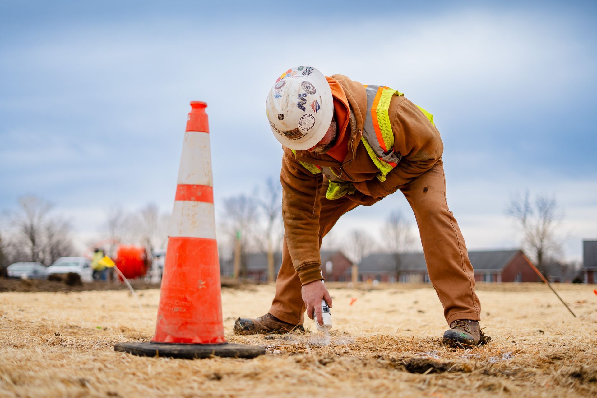 Construction worker in hard hat bending over a dirt lot beside an orange traffic cone, marking a spot