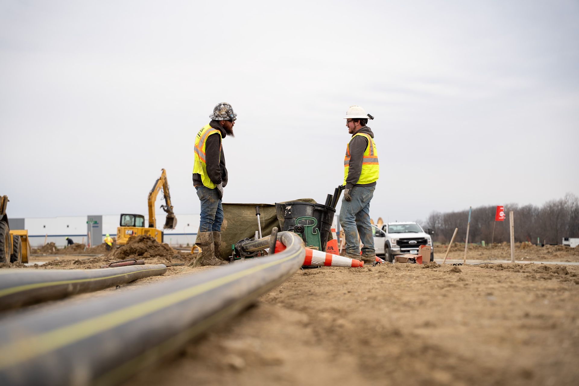 Two construction workers in safety vests stand on a dirt worksite with a road pipe in the foreground.