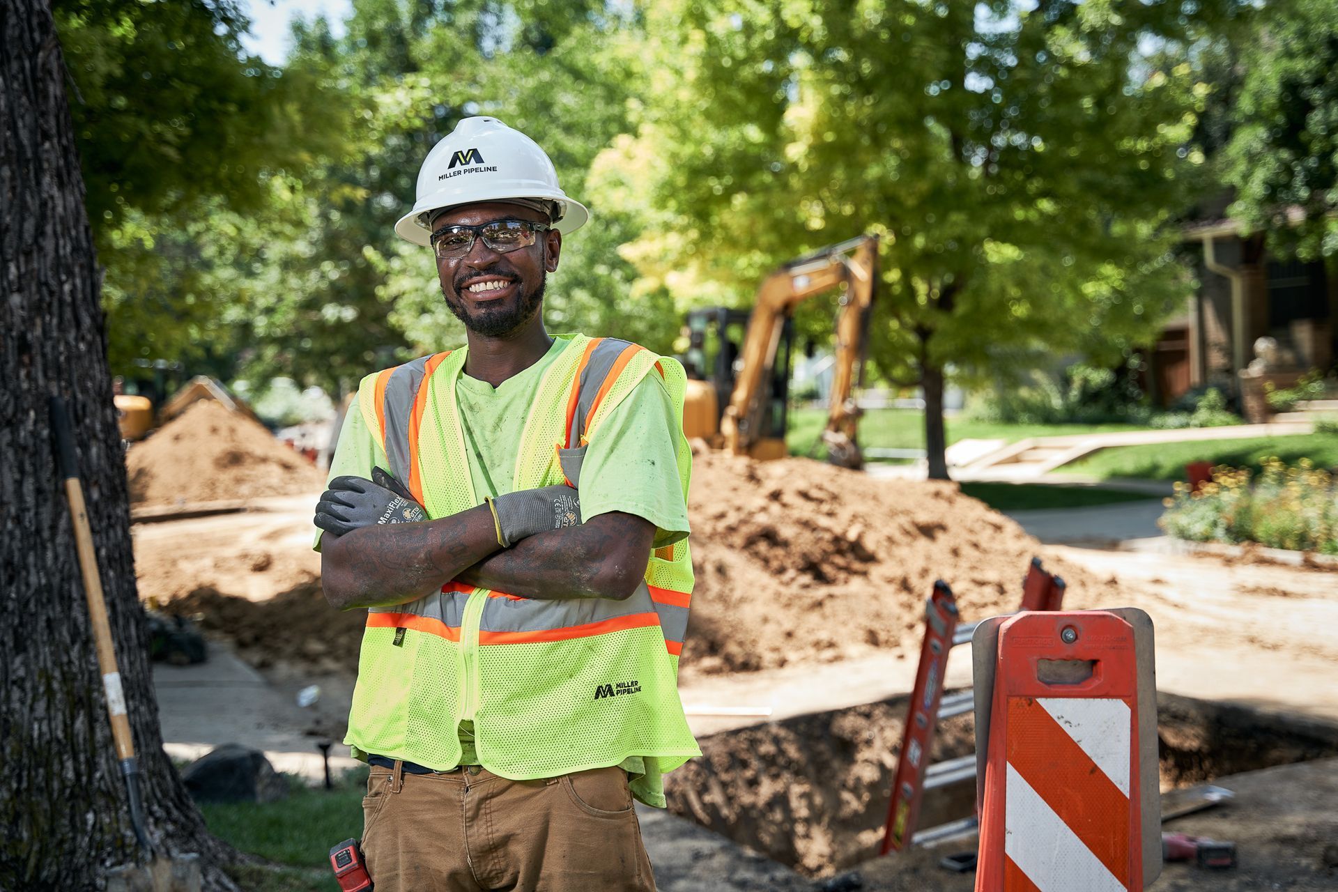 Construction worker in neon vest and hard hat stands with arms crossed at a roadwork site.