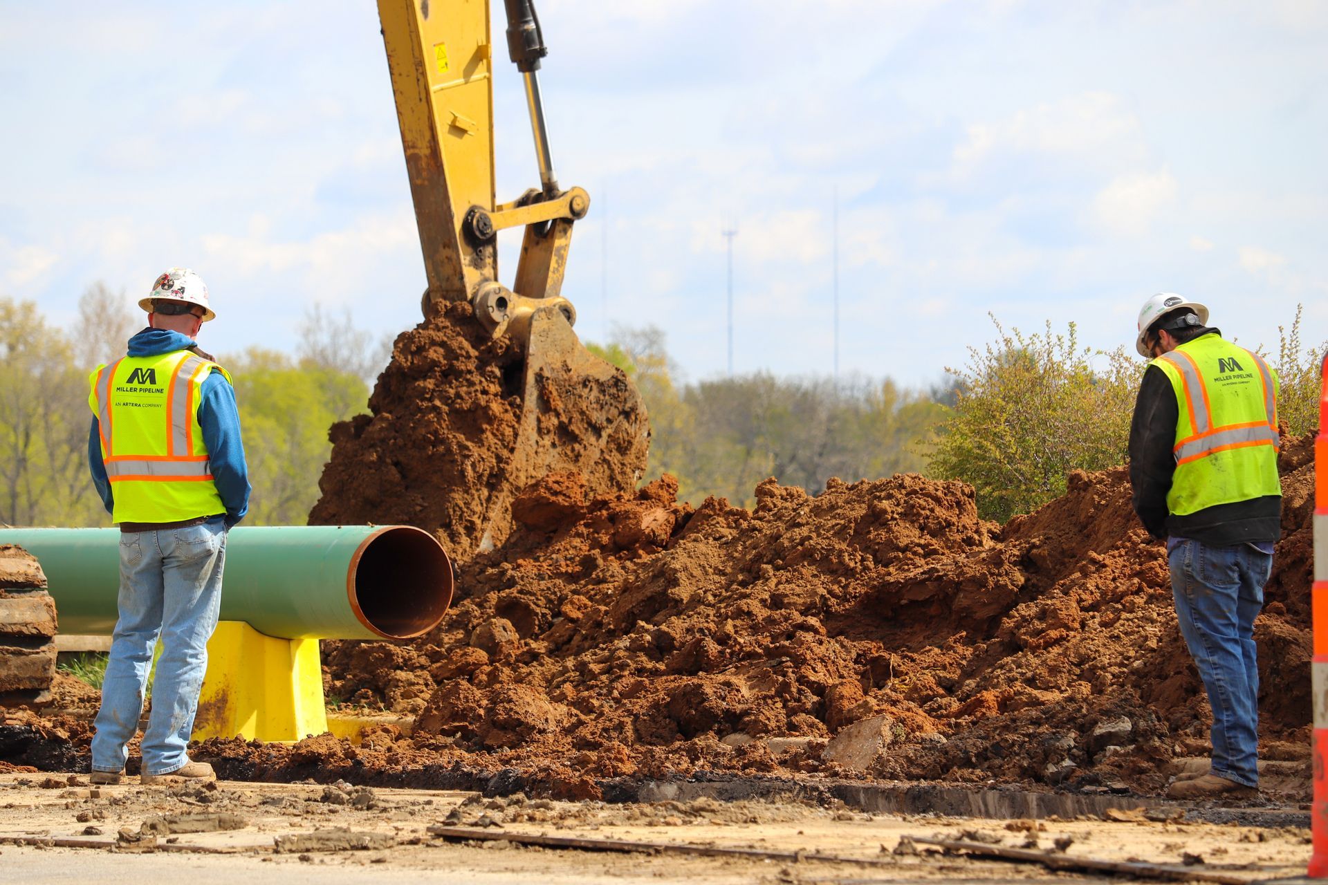Workers in safety vests dig around a large pipe with an excavator at a construction site