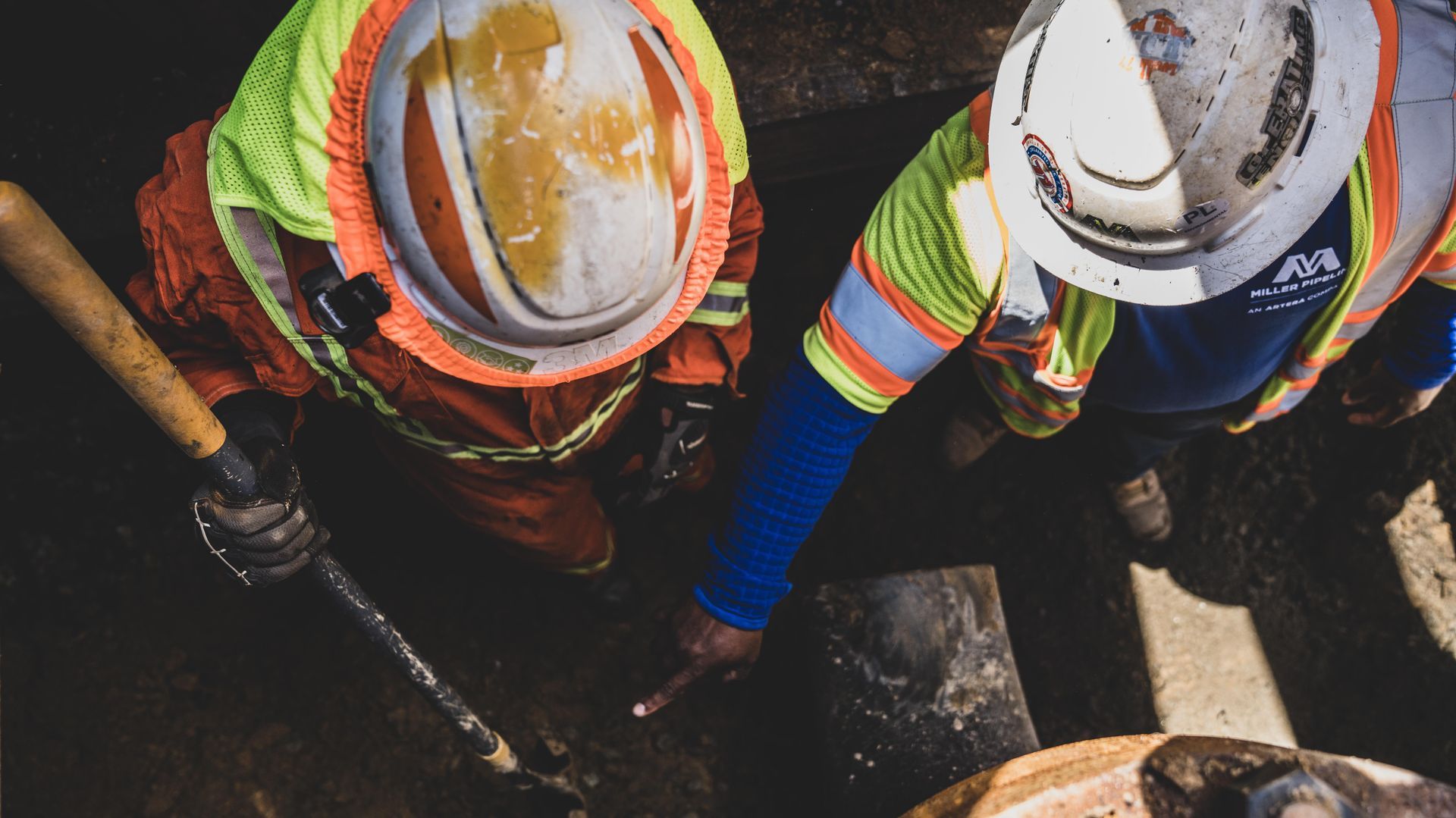 Two utility workers in hard hats and safety vests working with tools in a dark trench