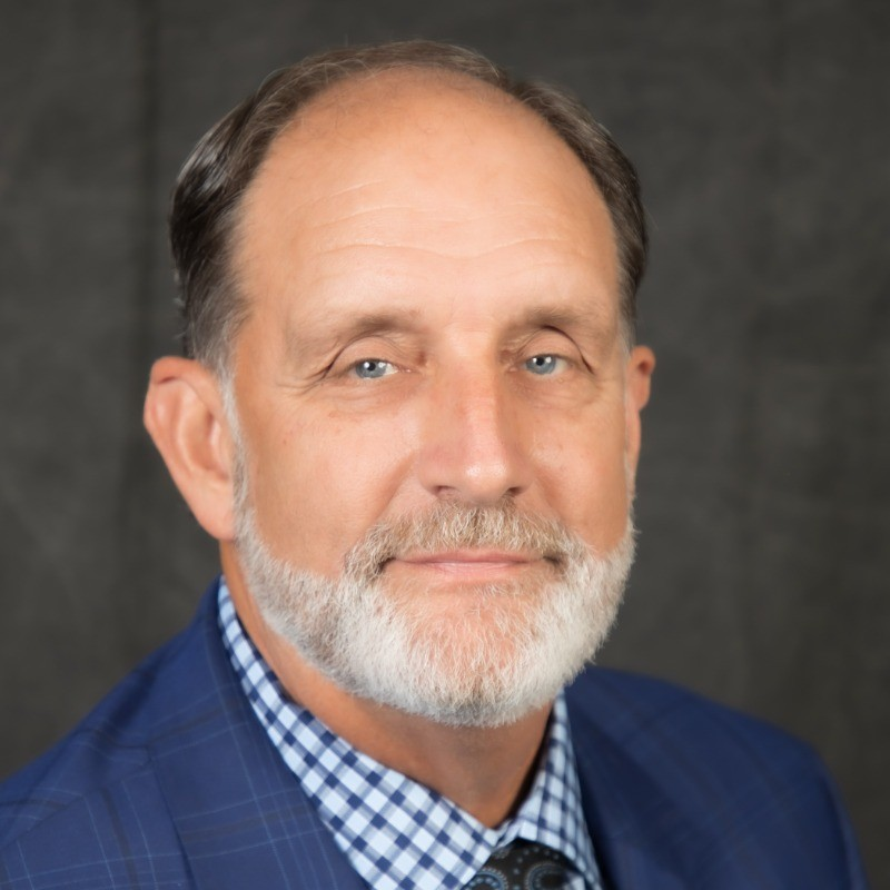 Studio headshot of a man in a blue suit and checkered shirt, smiling against a dark background