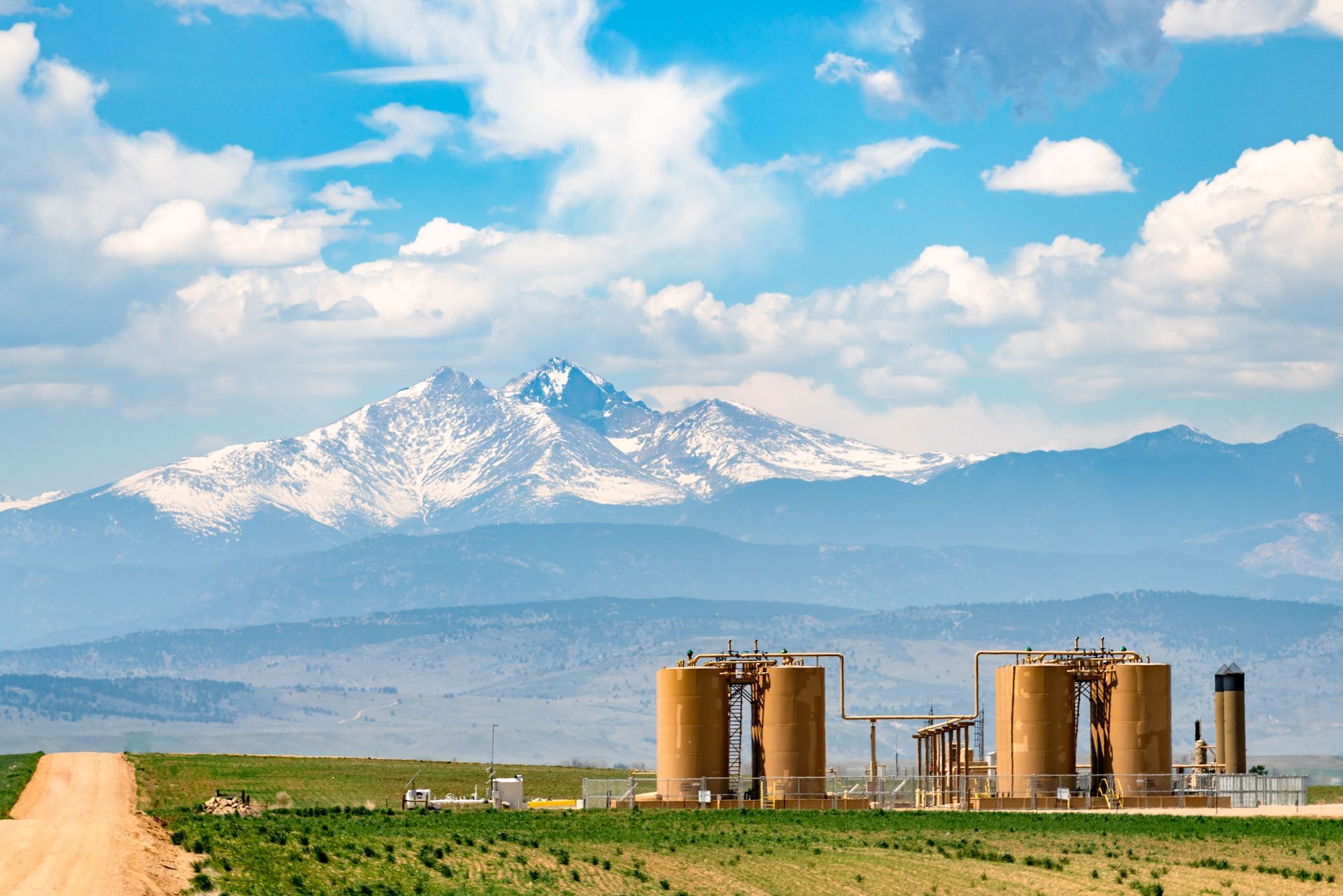 Snowy mountains under a blue sky above a grassy field and industrial tanks