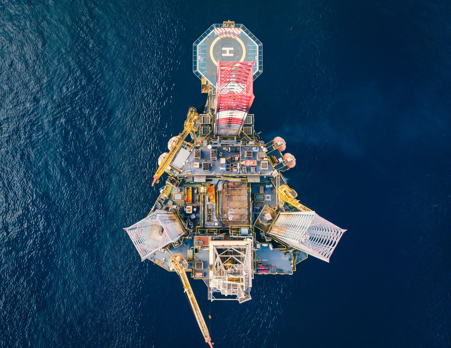 Aerial view of an offshore oil rig floating in deep blue water, with red and yellow structures visible.