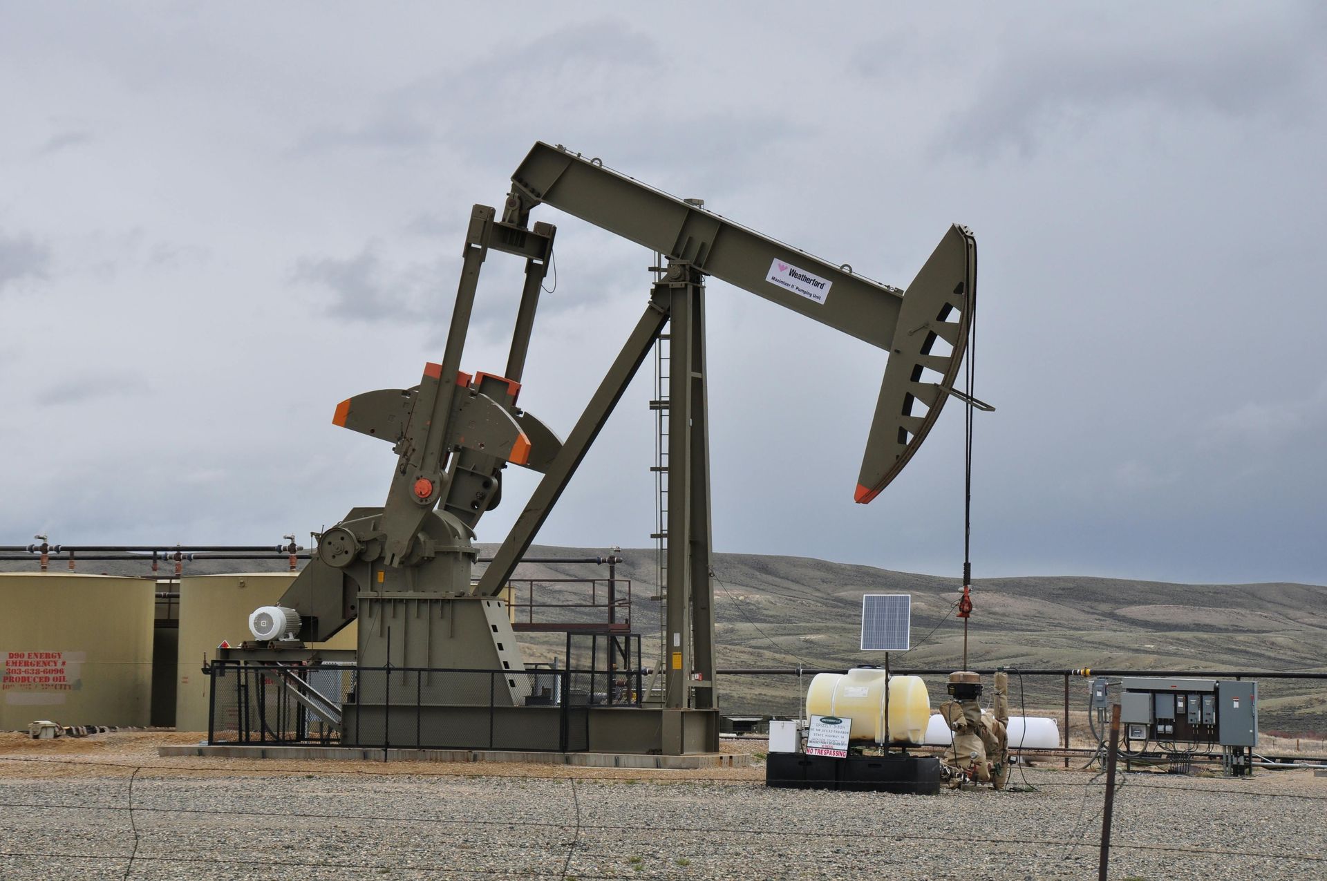 Oil pumpjack in a gravel field under a cloudy sky, with storage tanks and equipment nearby.