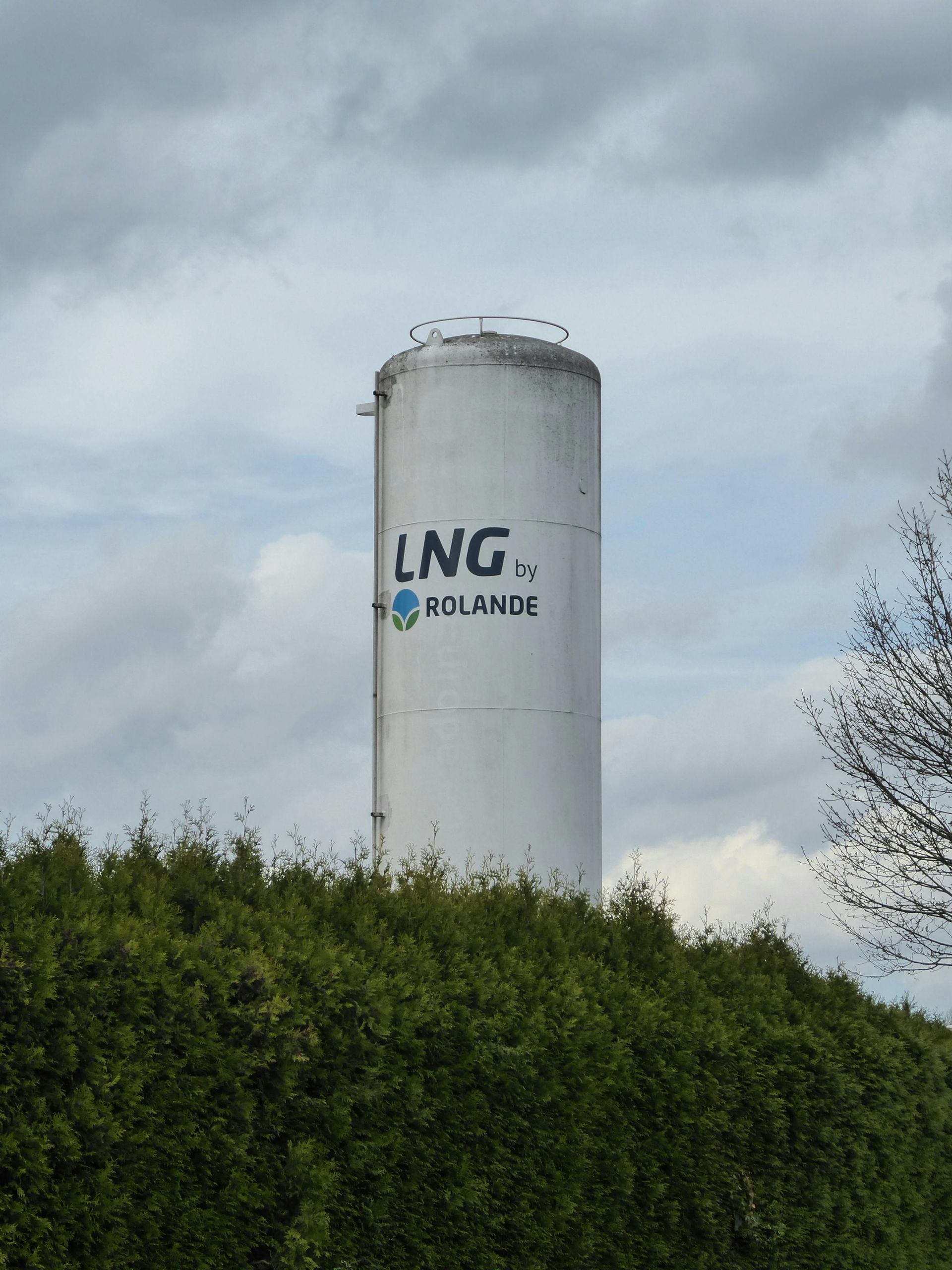 White industrial tank labeled “LNG” rising above green trees under a cloudy sky