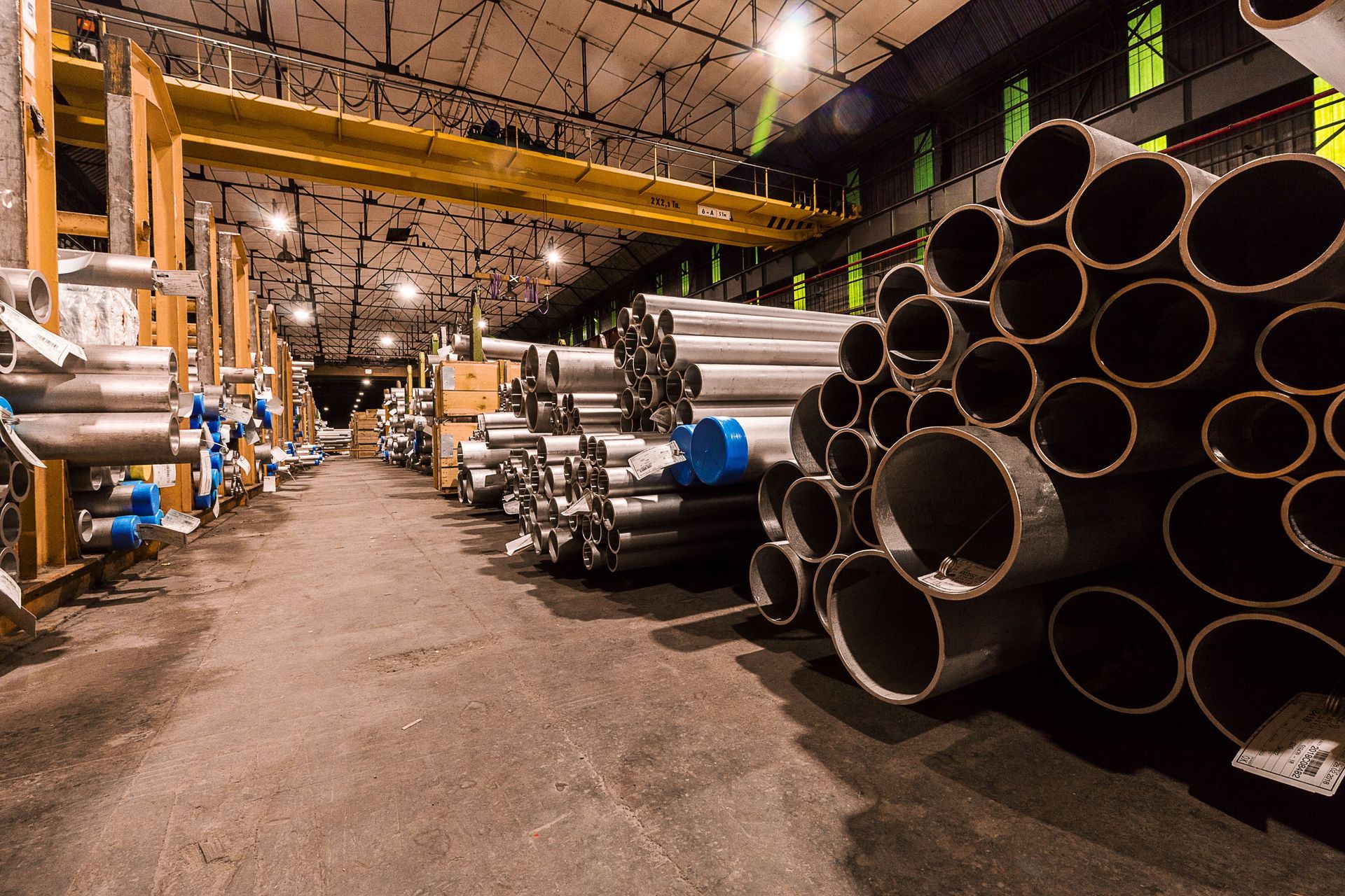 Industrial warehouse aisle with stacked metal pipes and tubing under overhead lights