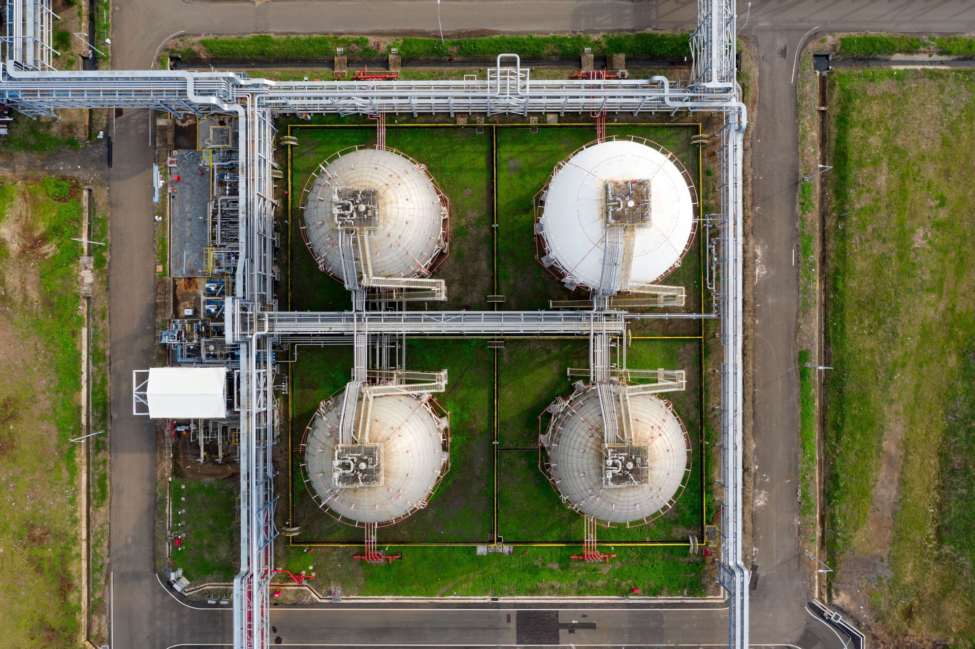 Aerial view of four white industrial storage tanks in a fenced facility with pipelines and grassy areas.