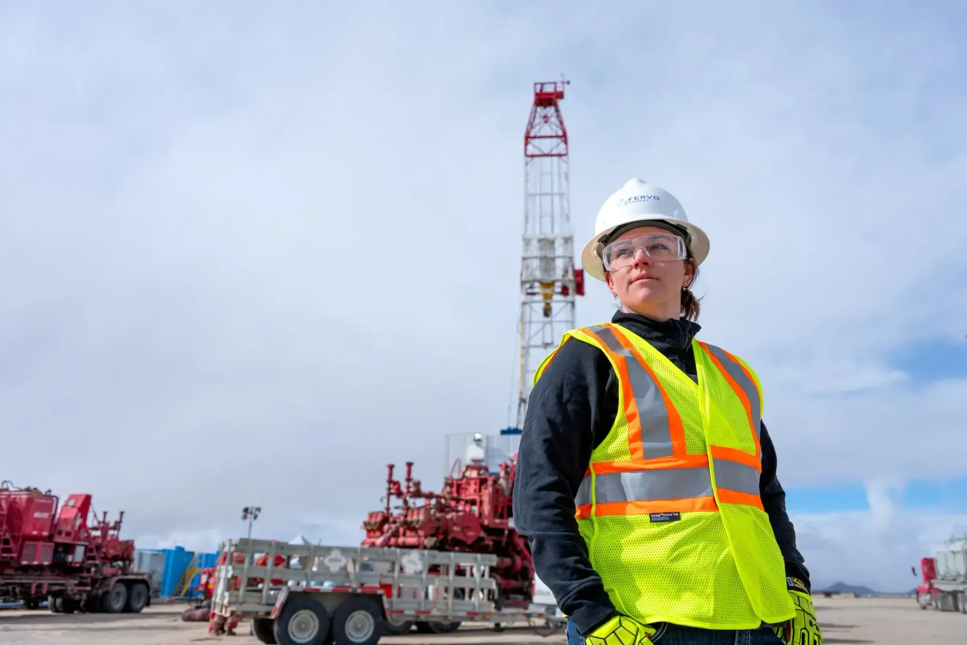 Construction worker in hard hat and orange vest at an industrial site with drilling equipment.