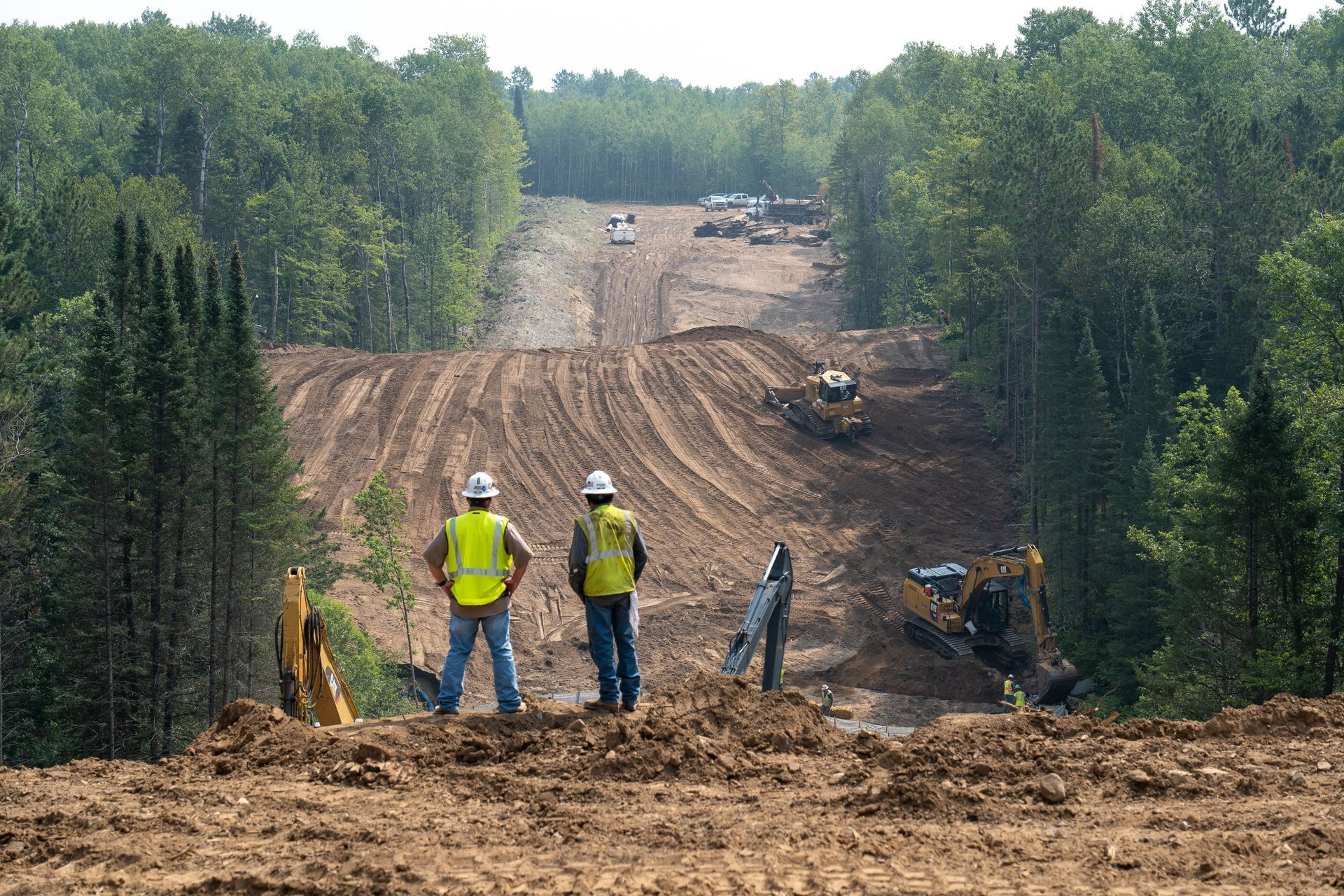 Construction workers in yellow vests overlook a dirt road cut through a forested hillside.