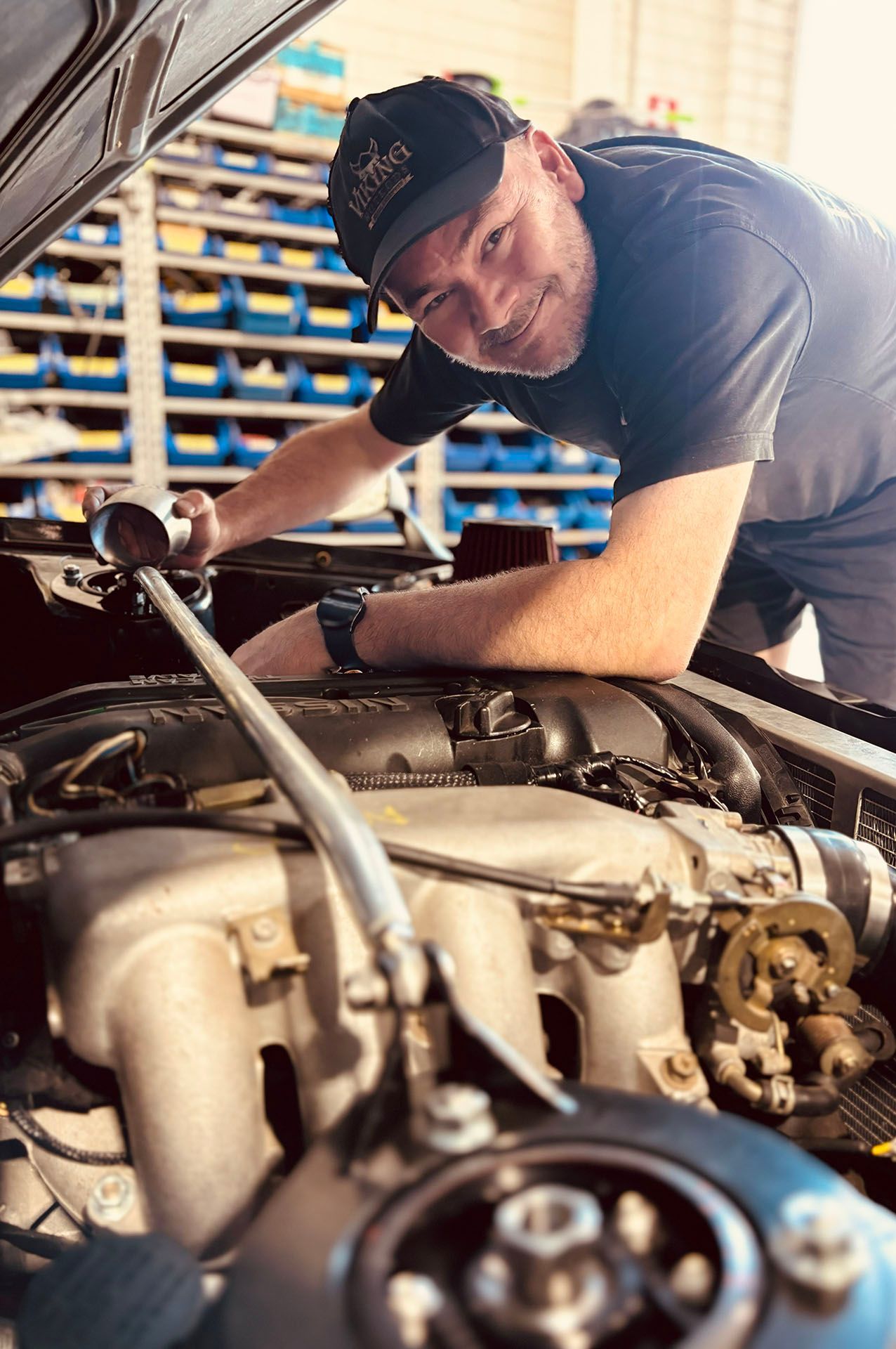 Mechanic leaning over an engine bay in a garage, working on a car with tools.