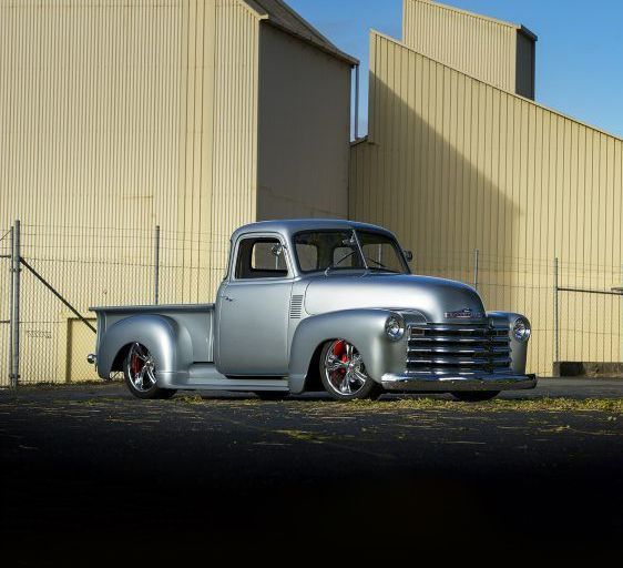 Silver custom vintage pickup truck parked beside a beige industrial building