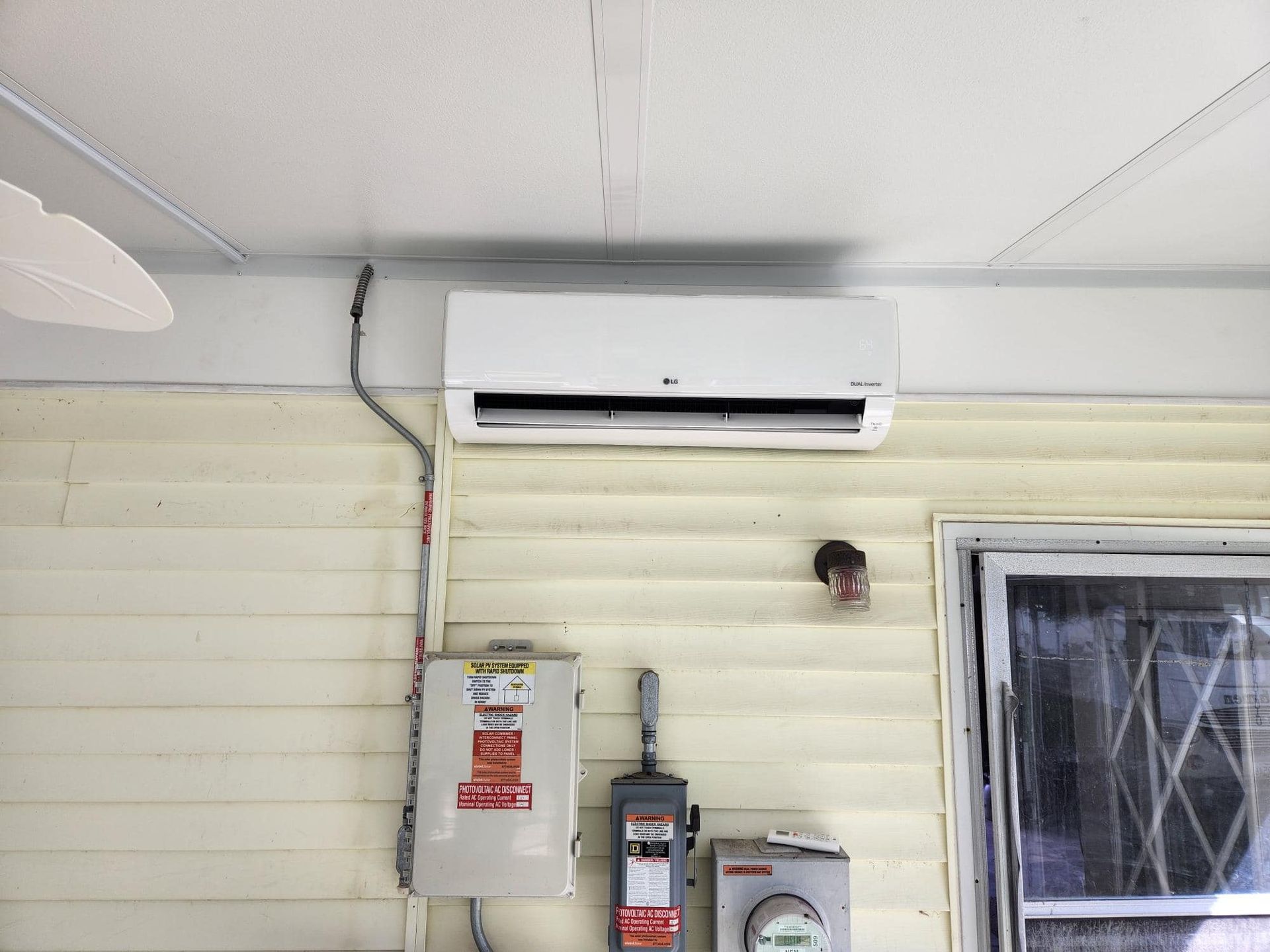 Wall-mounted air conditioner above electrical boxes on a beige exterior wall, with a window on the right.