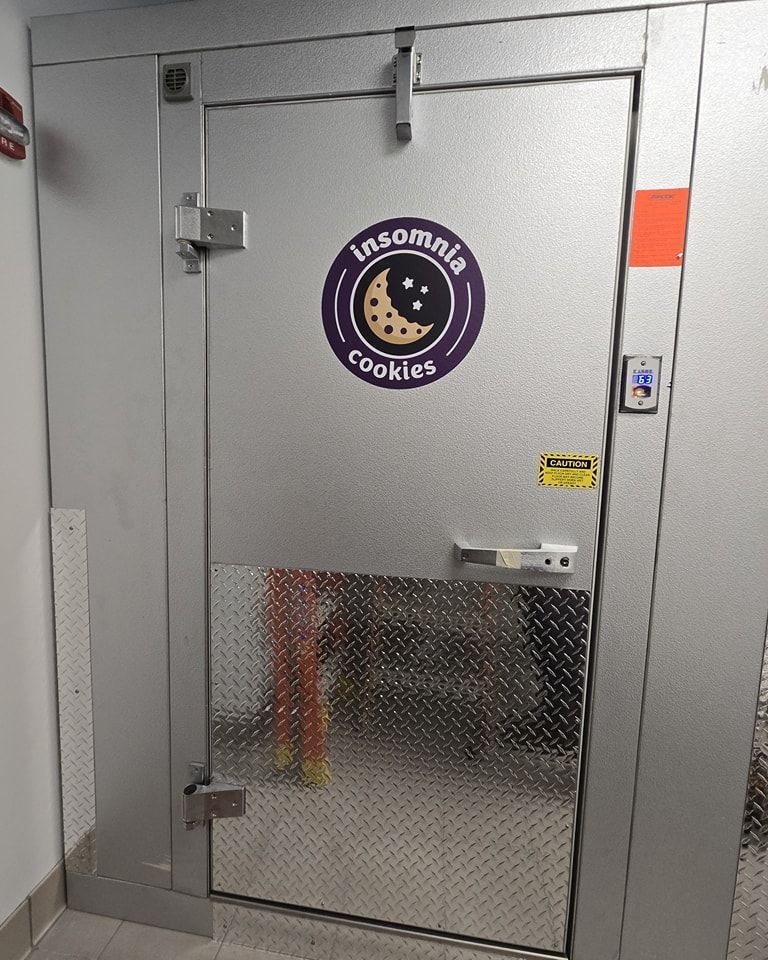 Gray metal walk-in freezer door with a “The Cookie Company” logo and a small keypad lock.